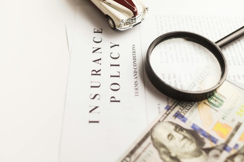 Close up of an insurance policy document with a magnifying glass, a toy car, and a hundred dollar bill on top.