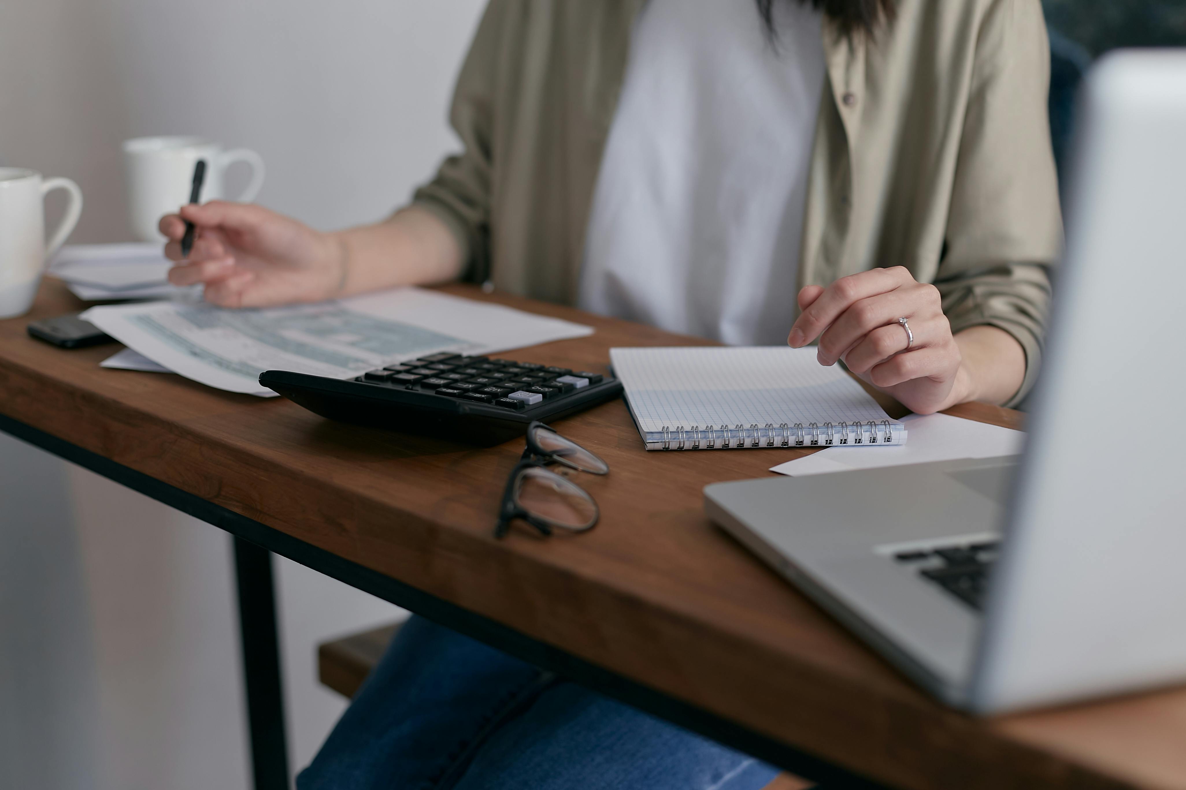 A person seated at a wooden desk using a calculator and taking notes, with a laptop, glasses, and coffee nearby
