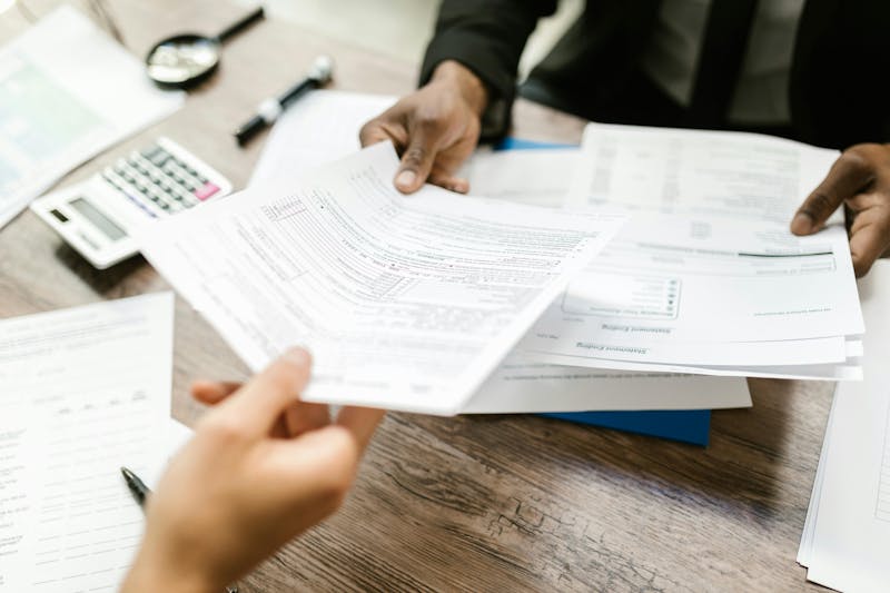 Two people are looking at and handing each other paperwork on a desk with a calculator and pen nearby.