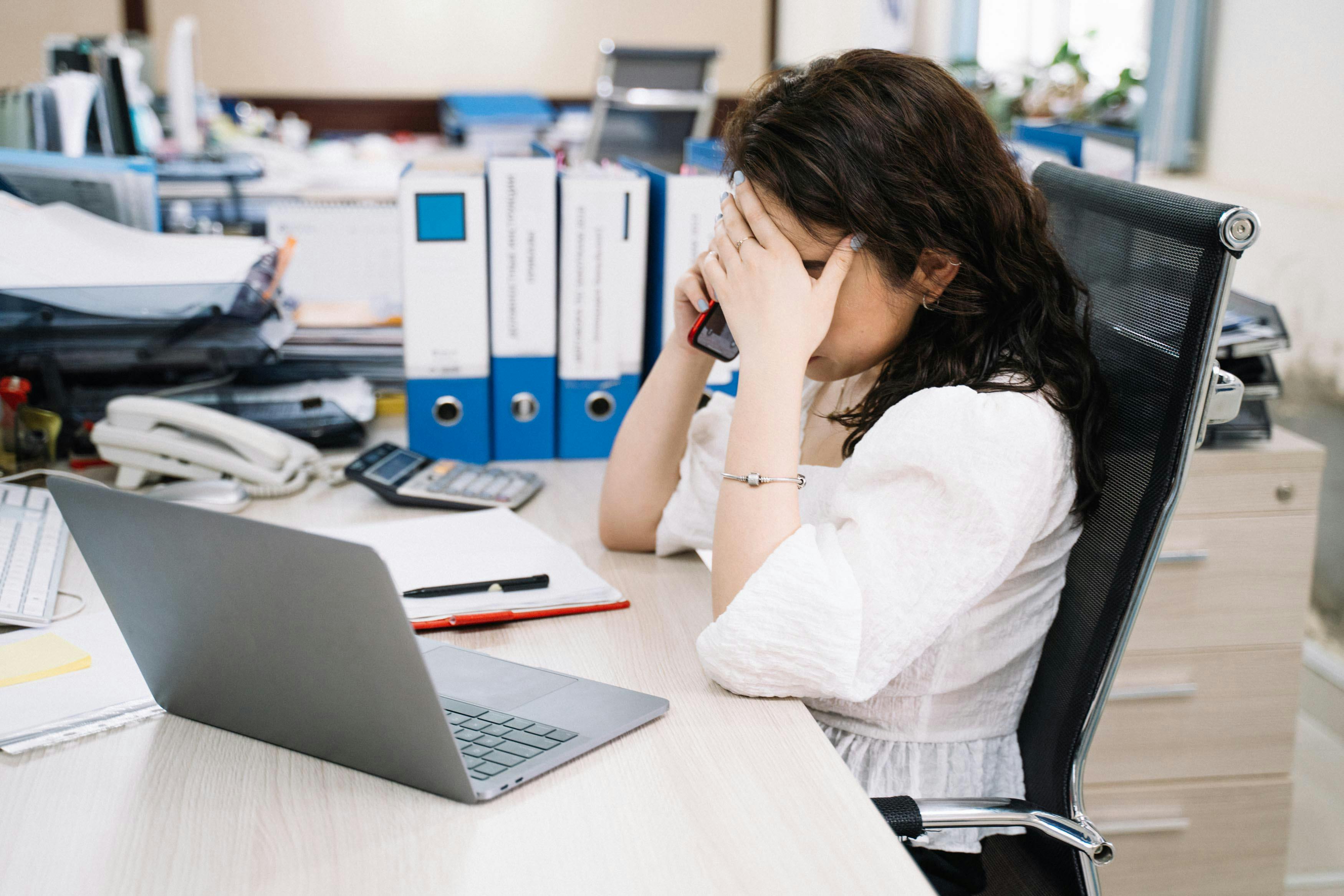 A woman is sitting at a messy desk, holding her head in one hand and a phone in the other, looking stressed or overwhelmed.