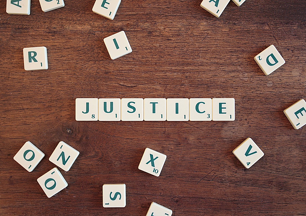 The word "JUSTICE" is spelled out using letter tiles on a wooden table, with other scattered tiles around it.