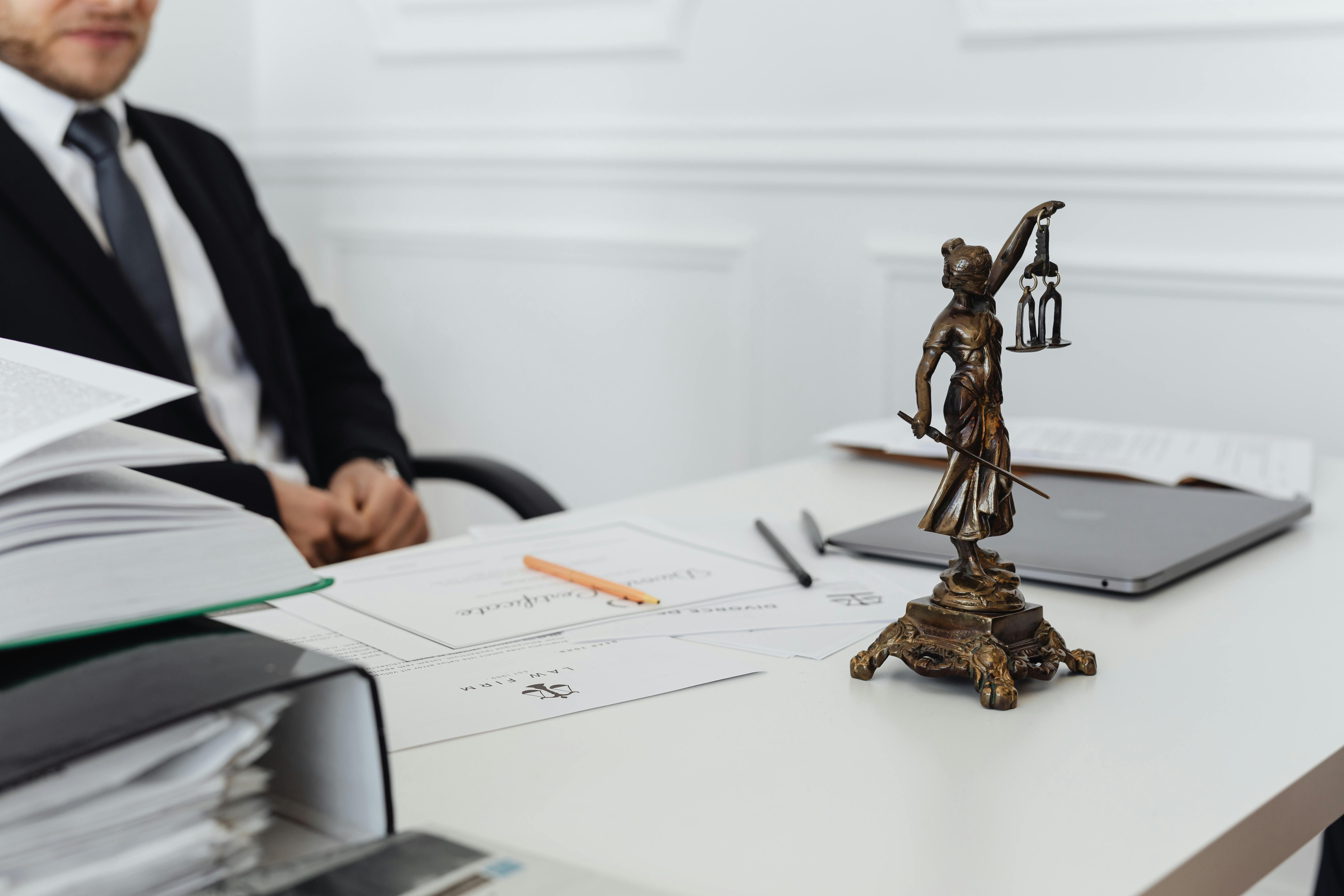 Close-up of a Justice statue on a lawyer’s desk, with legal documents, a laptop, and a person in a suit sitting in the background.