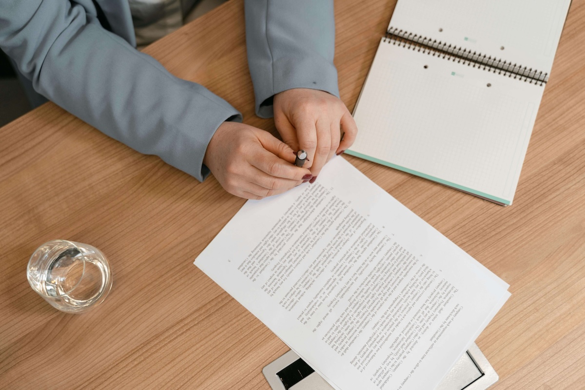 A person in a blue blazer holding a pen while reviewing a document on a wooden desk with a notebook and a glass of water nearby.