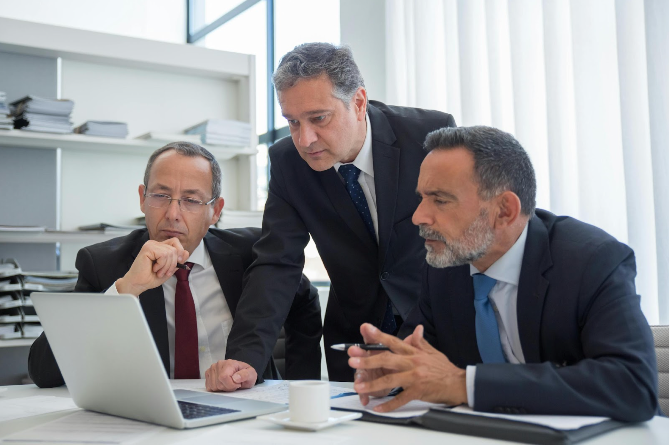 Three businessmen in suits are gathered around a laptop, focused on the screen during a meeting.