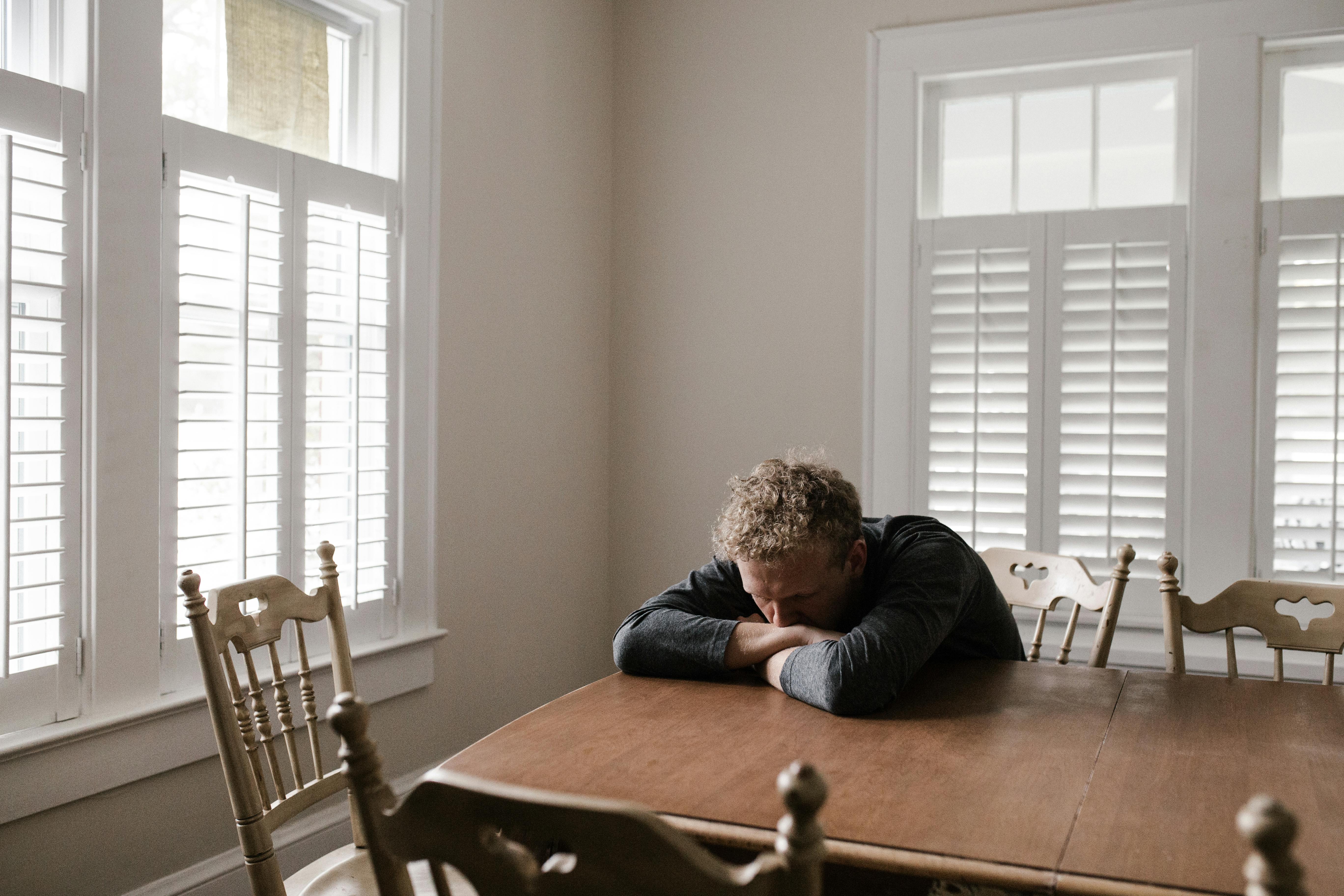 Man sitting at a wooden table with his head resting on his arms, appearing sad or tired in a bright room.