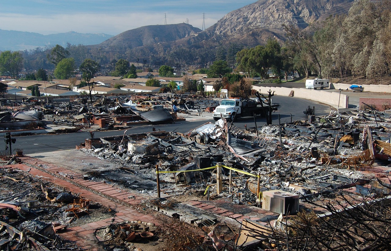 Widespread destruction in a residential area after a wildfire, with charred remains of homes and vehicles, and mountains visible in the background.