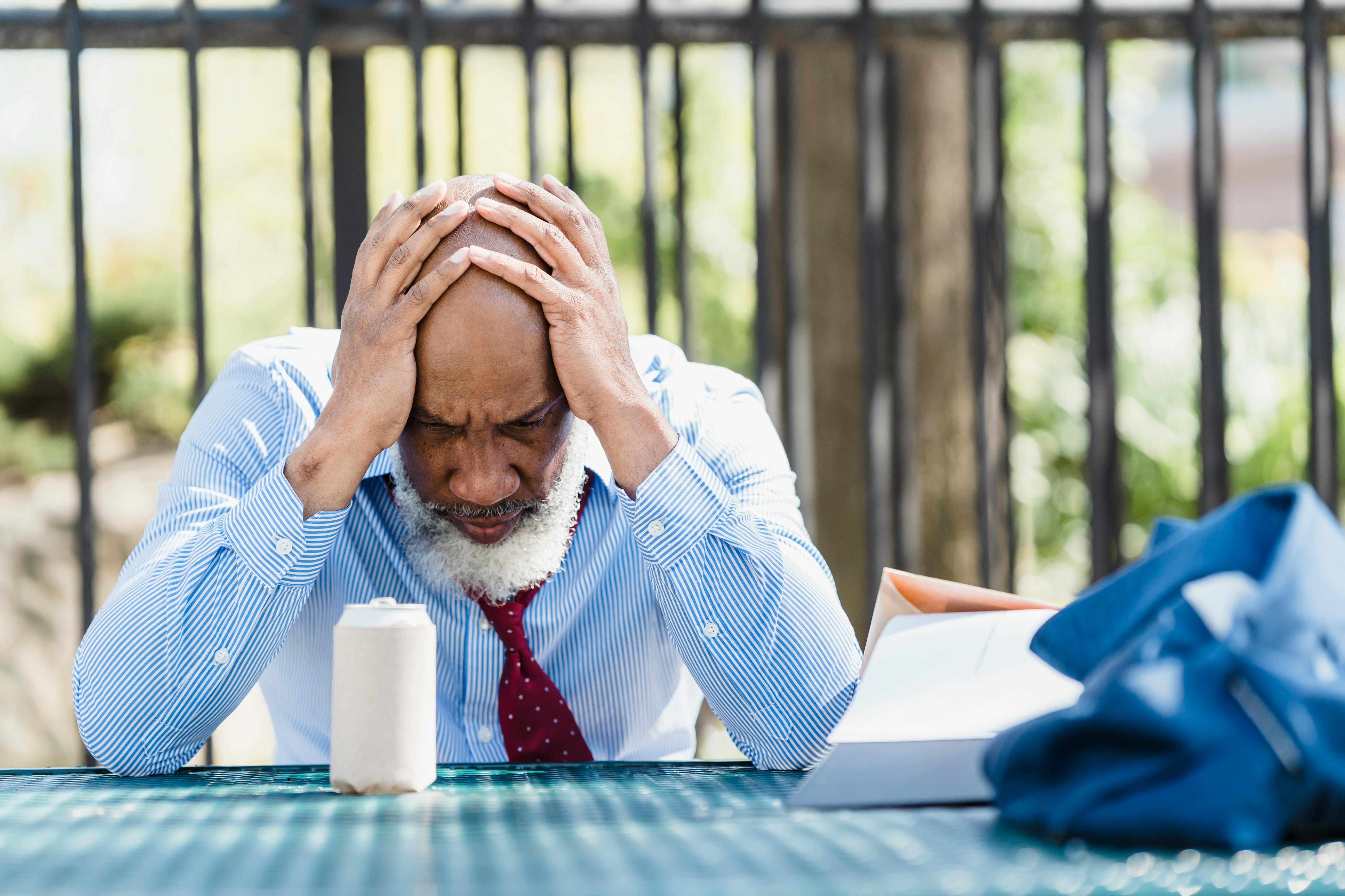 Stressed man sitting at a table outdoors with his head in his hands, surrounded by paperwork and a drink can, wearing a shirt and tie.