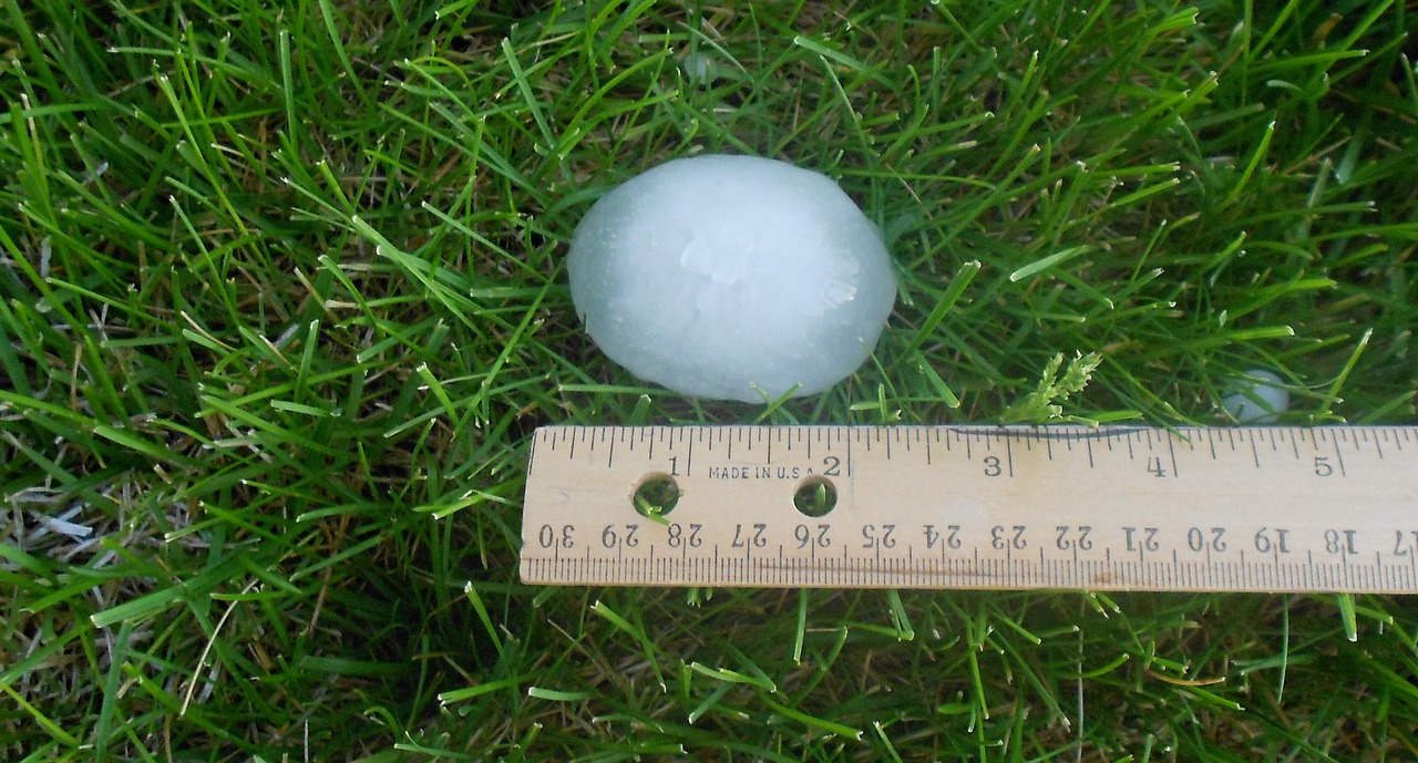 A large hailstone sits on grass next to a wooden ruler, showing it measures approximately 2.5 inches in diameter.