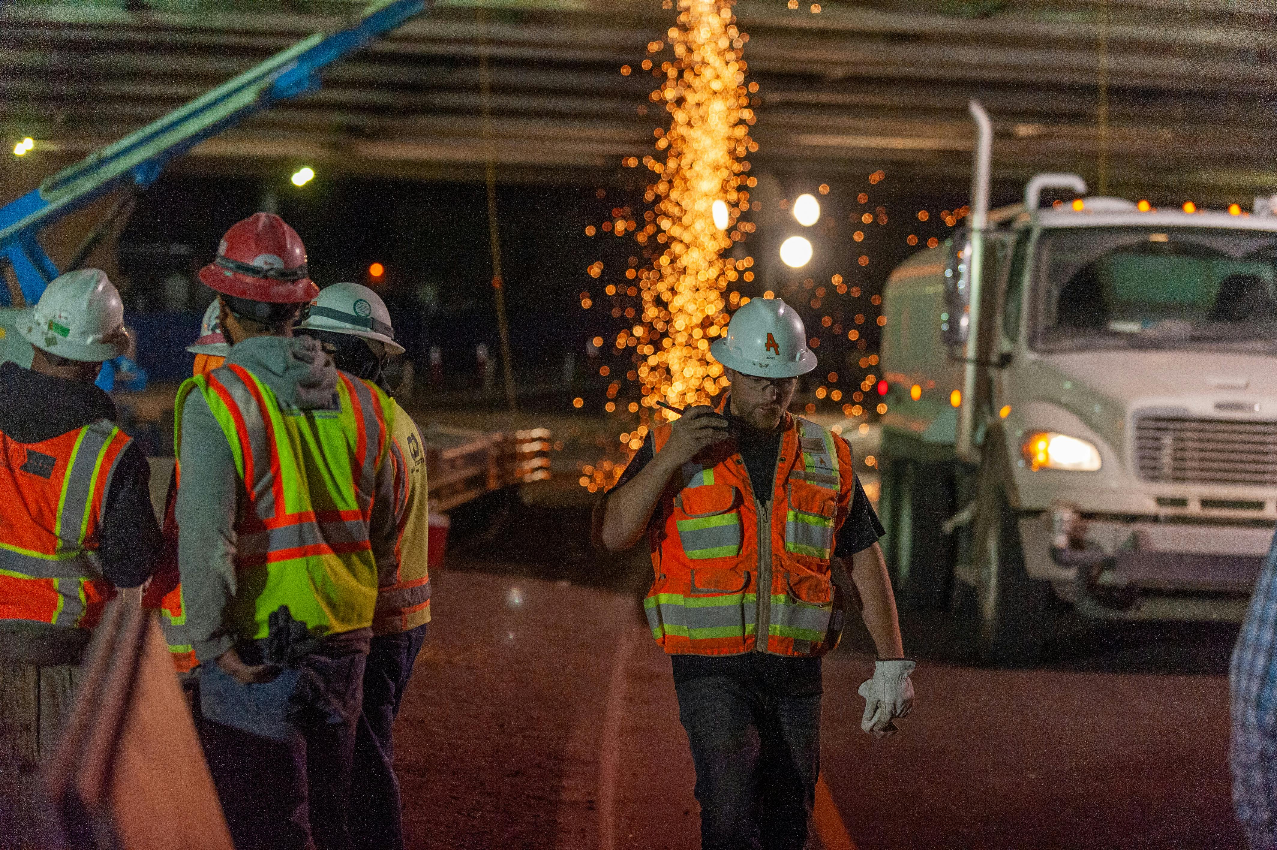 Construction workers in safety gear at a nighttime work site with sparks falling in the background.