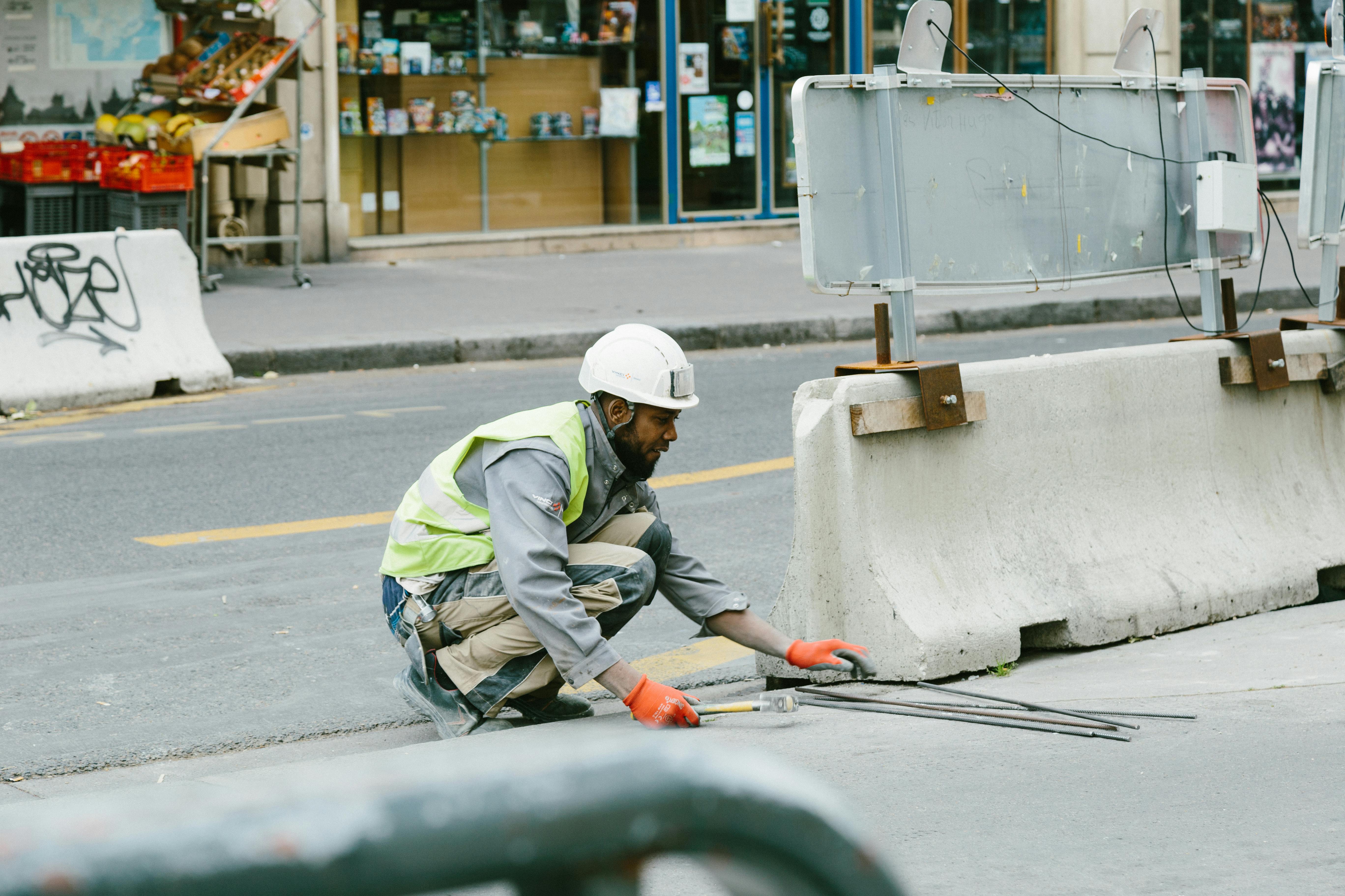 Construction worker in safety gear repairing pavement near a road barrier in an urban area.