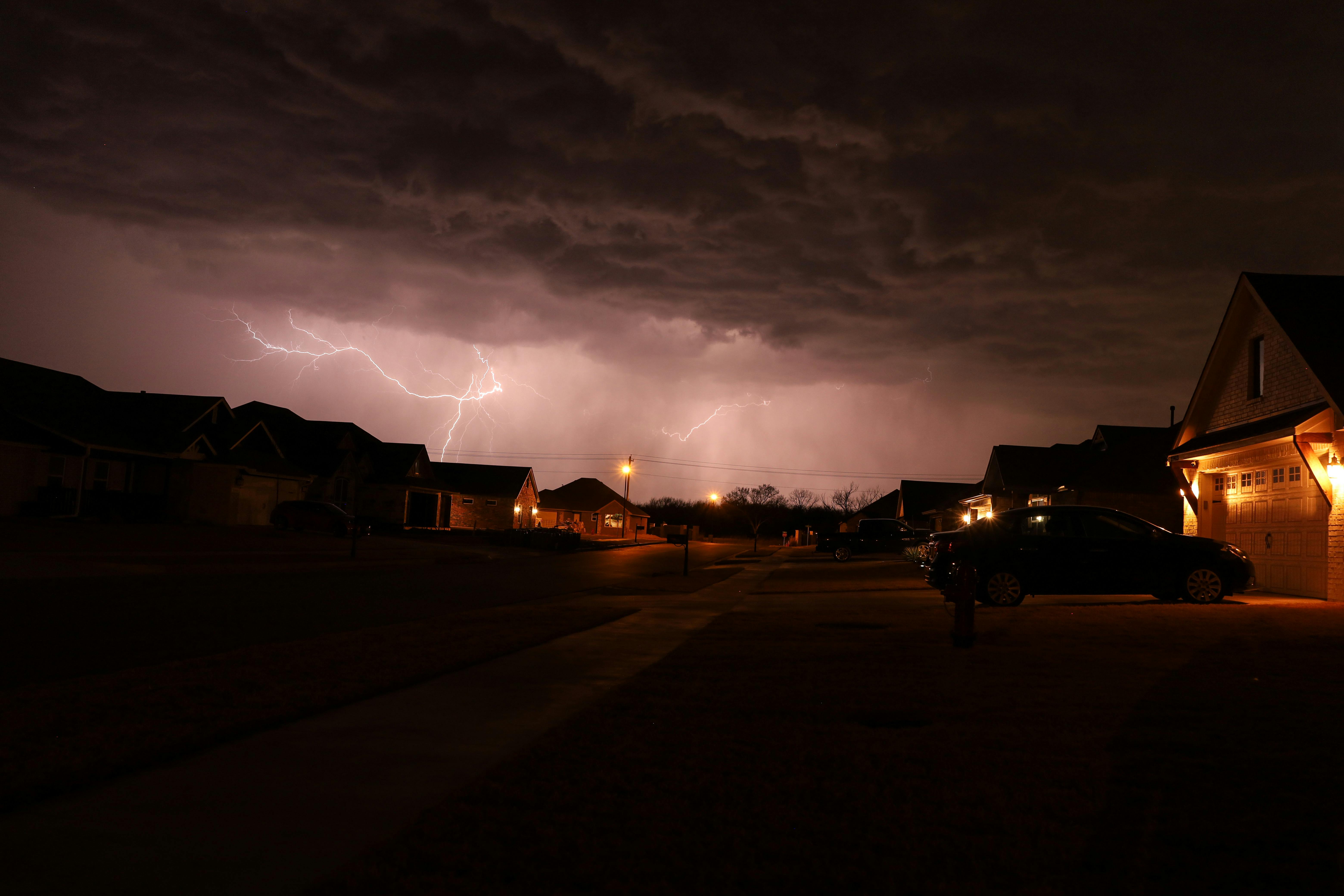 Lightning flashes across the sky above a quiet suburban street at night, with houses and parked cars illuminated by streetlights.