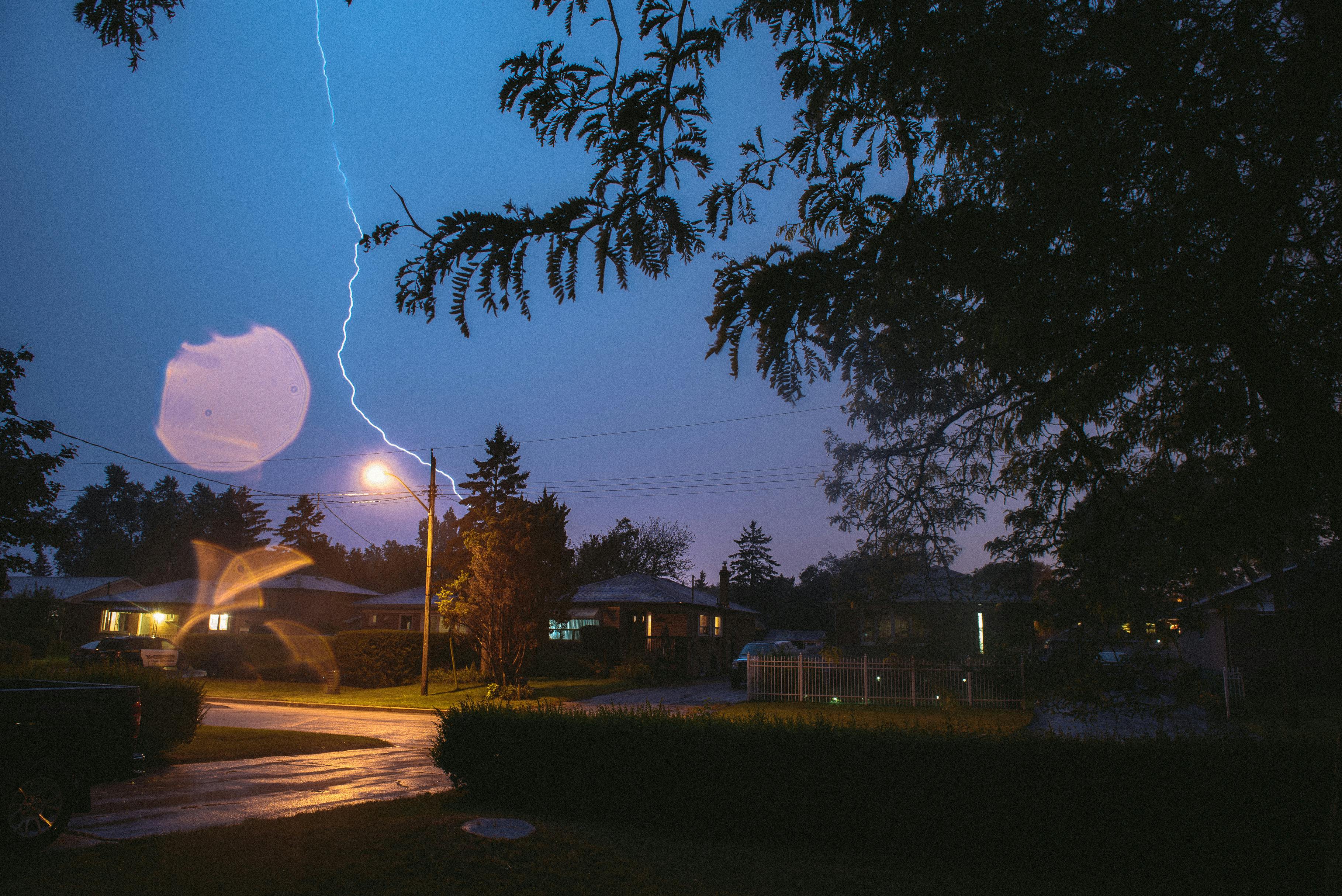 Lightning illuminates dark storm clouds above a suburban neighborhood with houses and trees.