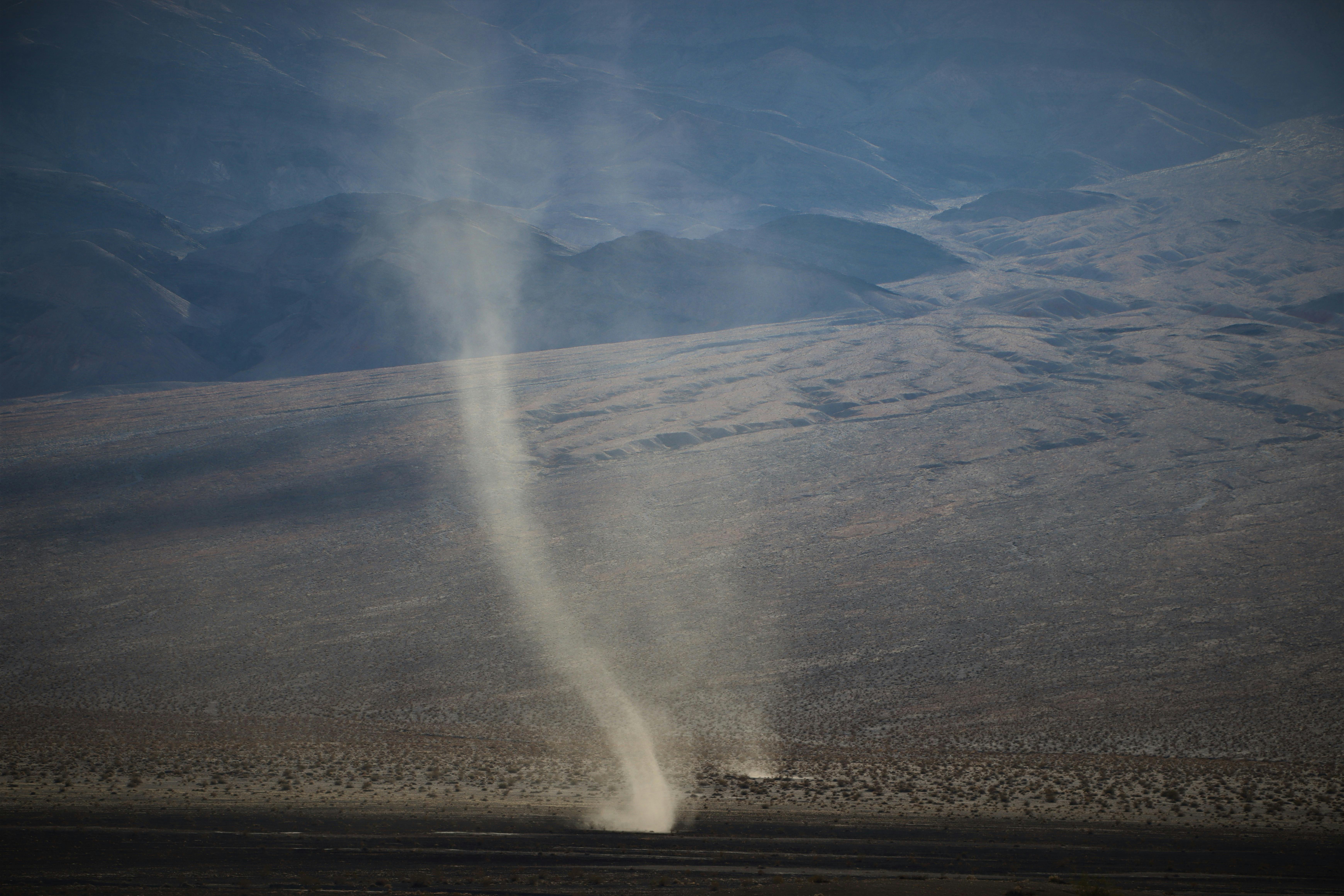 Dust devil swirling across a dry desert landscape with mountains in the background.