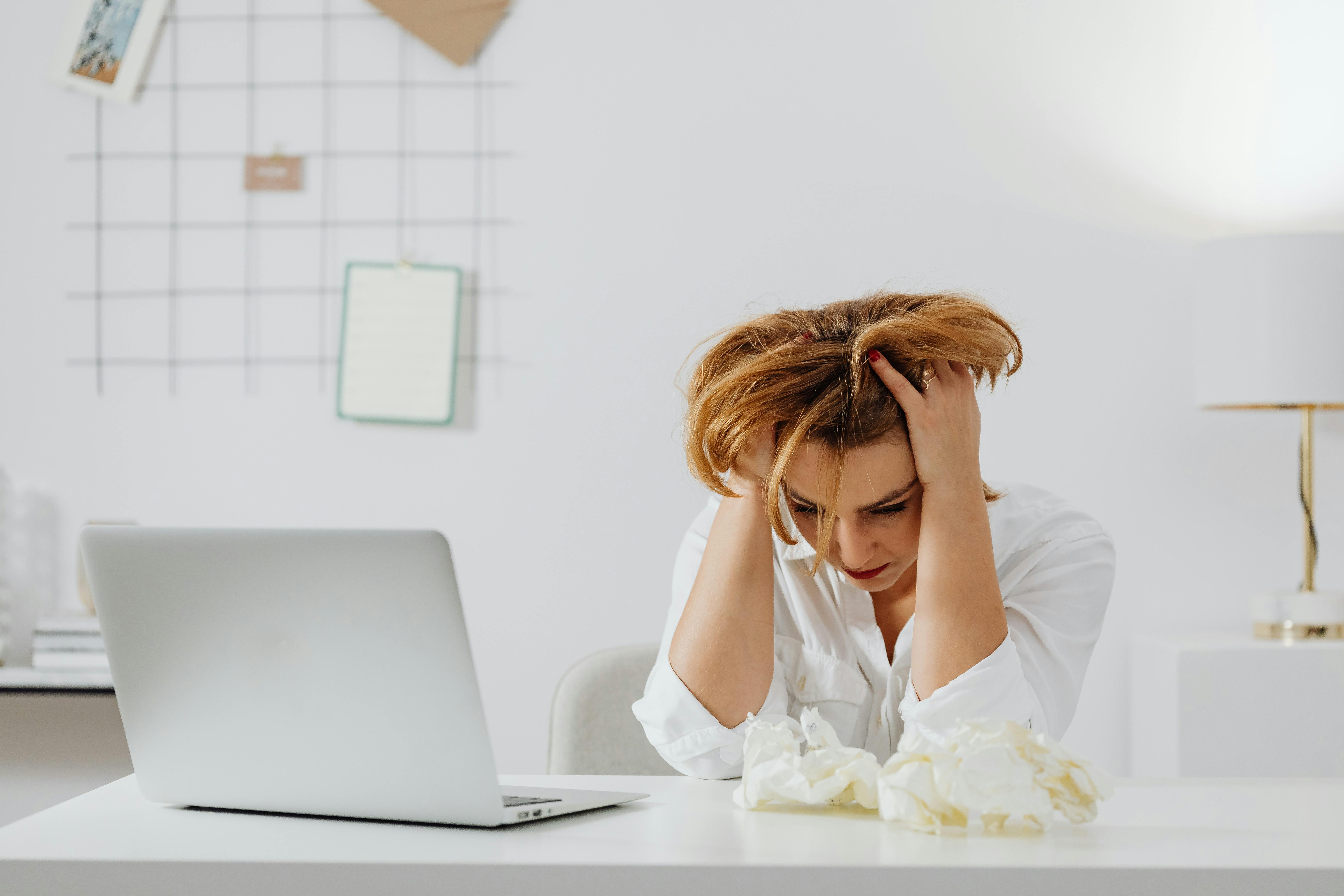 Frustrated woman sitting at a desk with her head in her hands, next to a laptop and crumpled tissues.