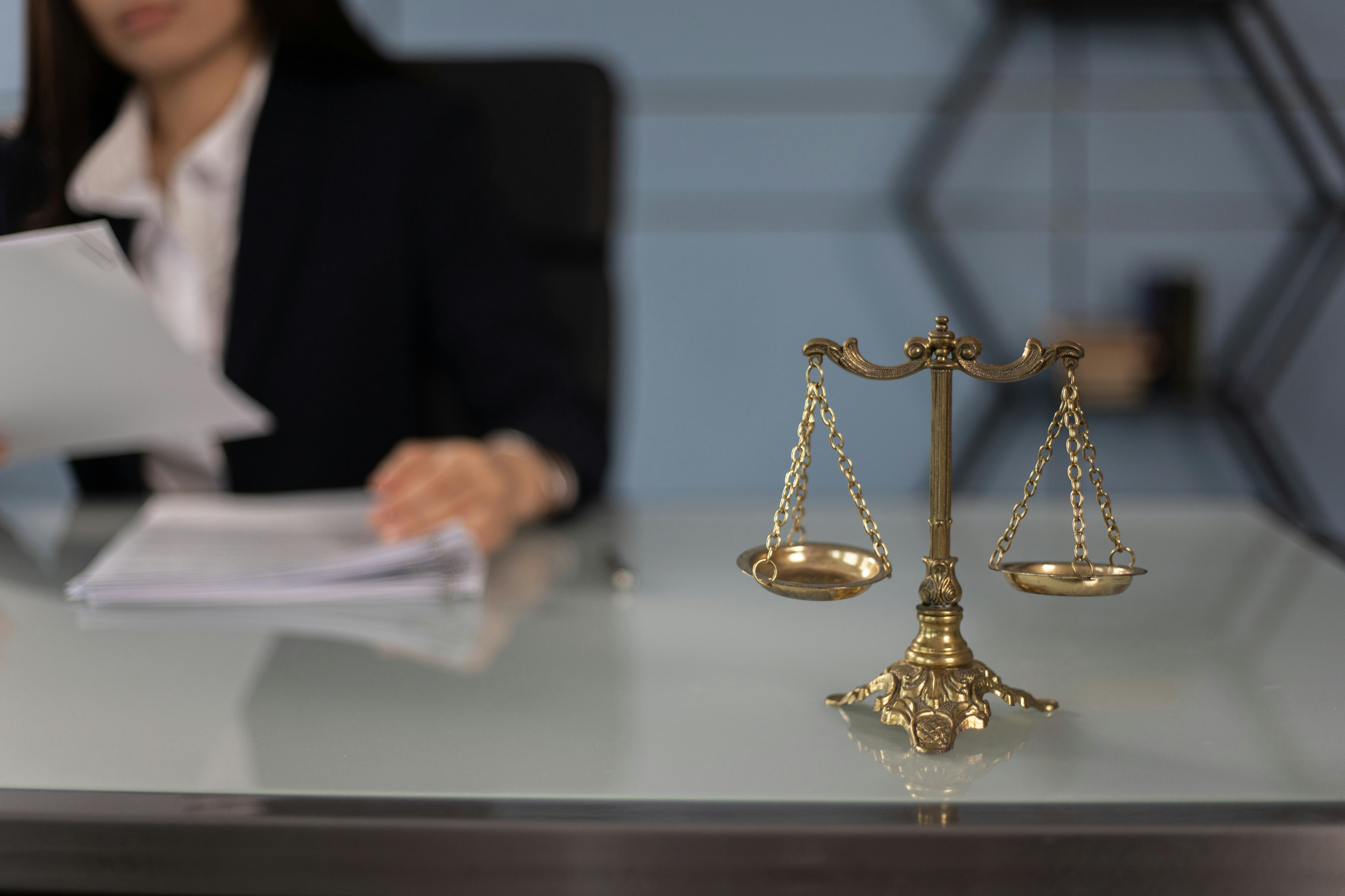 Golden balance scale on a desk with a lawyer holding documents in the background.