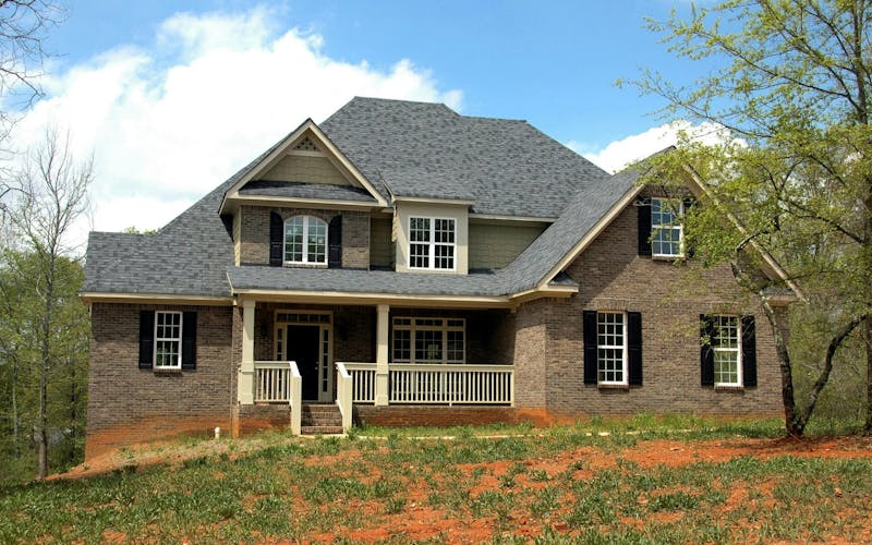 Two-story brick house with gray roof, white trim, and front porch surrounded by grass and trees.