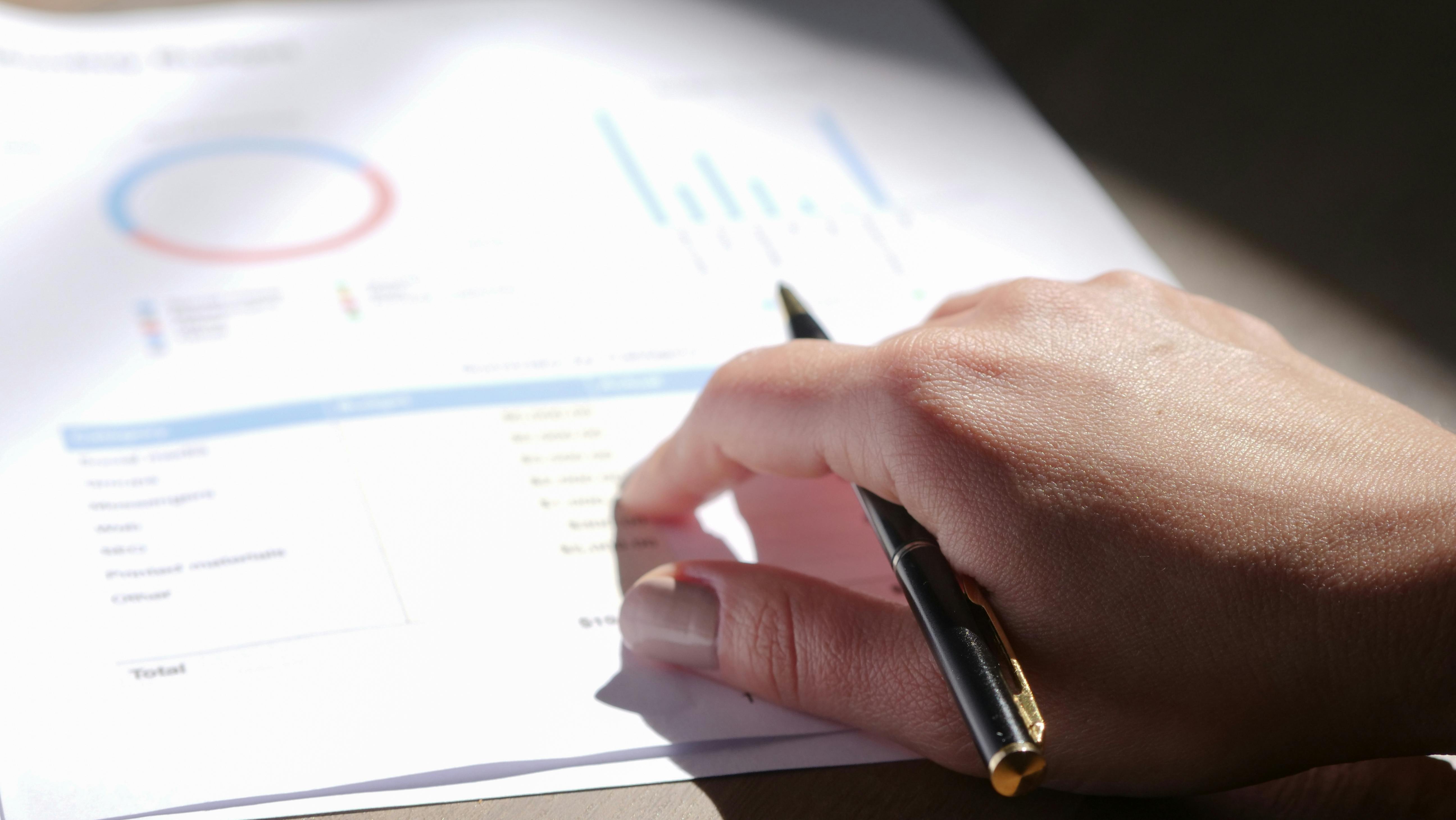 Hand holding a pen and pointing at a financial document with charts and graphs.
