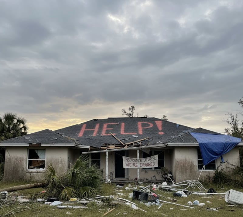 A damaged house with debris scattered outside, “HELP!” painted in large red letters on the roof, and a banner reading “Insurance Delayed, We’re Drained!” hanging over the front porch.