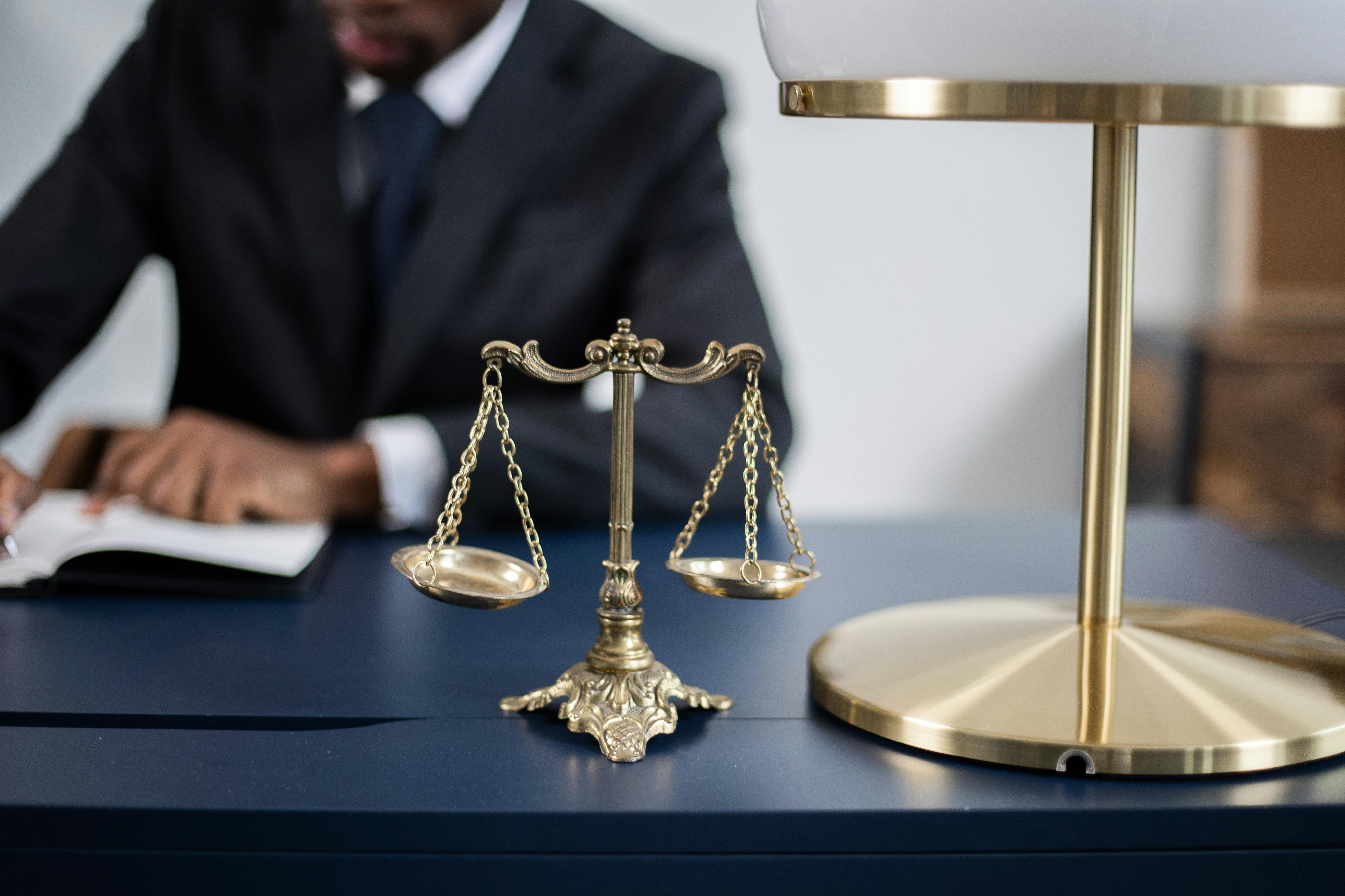 A brass balance scale on a desk with a person in a suit writing in the background.