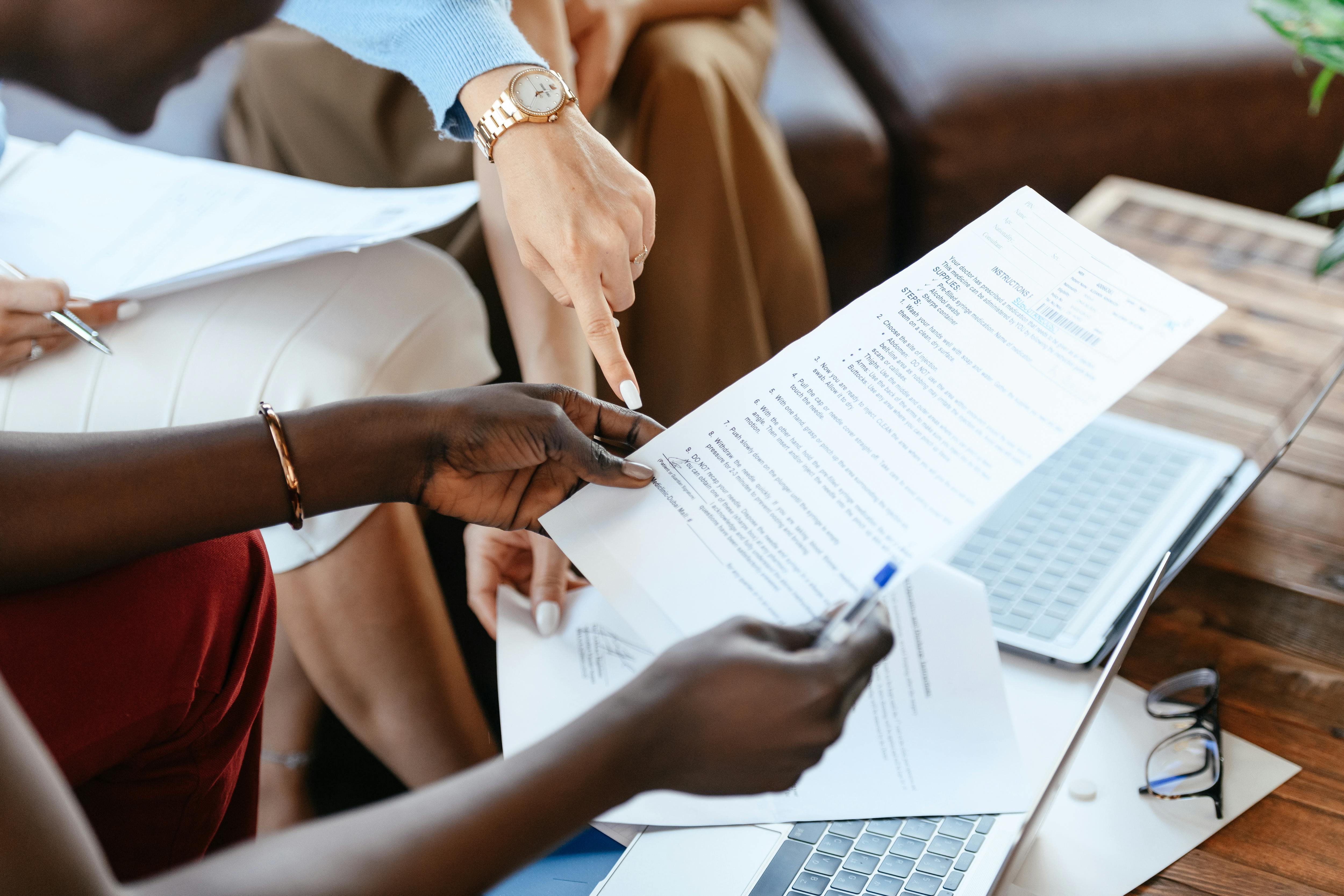 People collaborating on a document at a table, with one person pointing at a printed page while another holds it over a laptop.