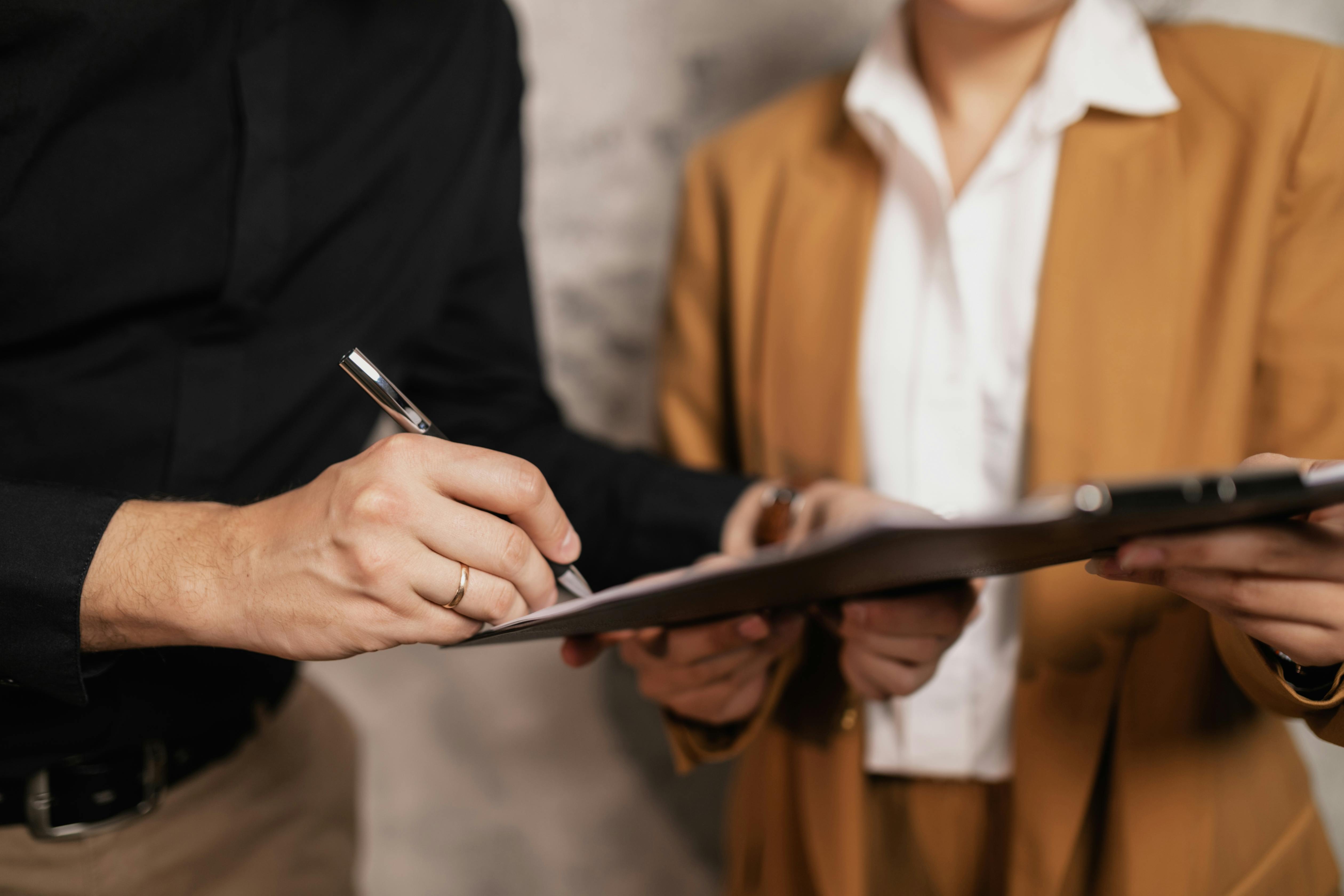 A person in a black shirt signs a document on a clipboard held by another person wearing a tan suit, suggesting a business agreement or contract review.