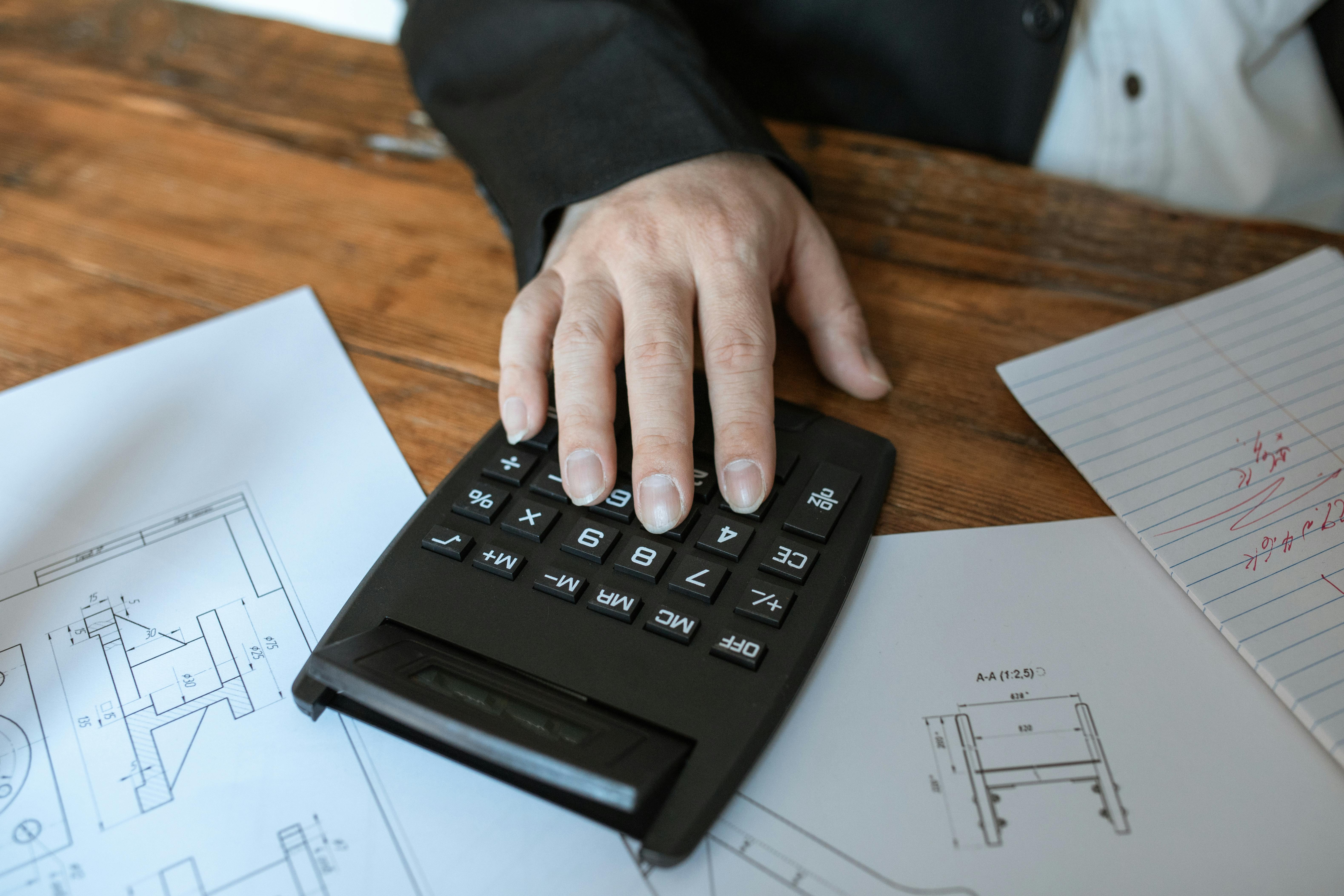 A person using a calculator on a wooden desk with engineering drawings and notes, calculating project or construction costs.