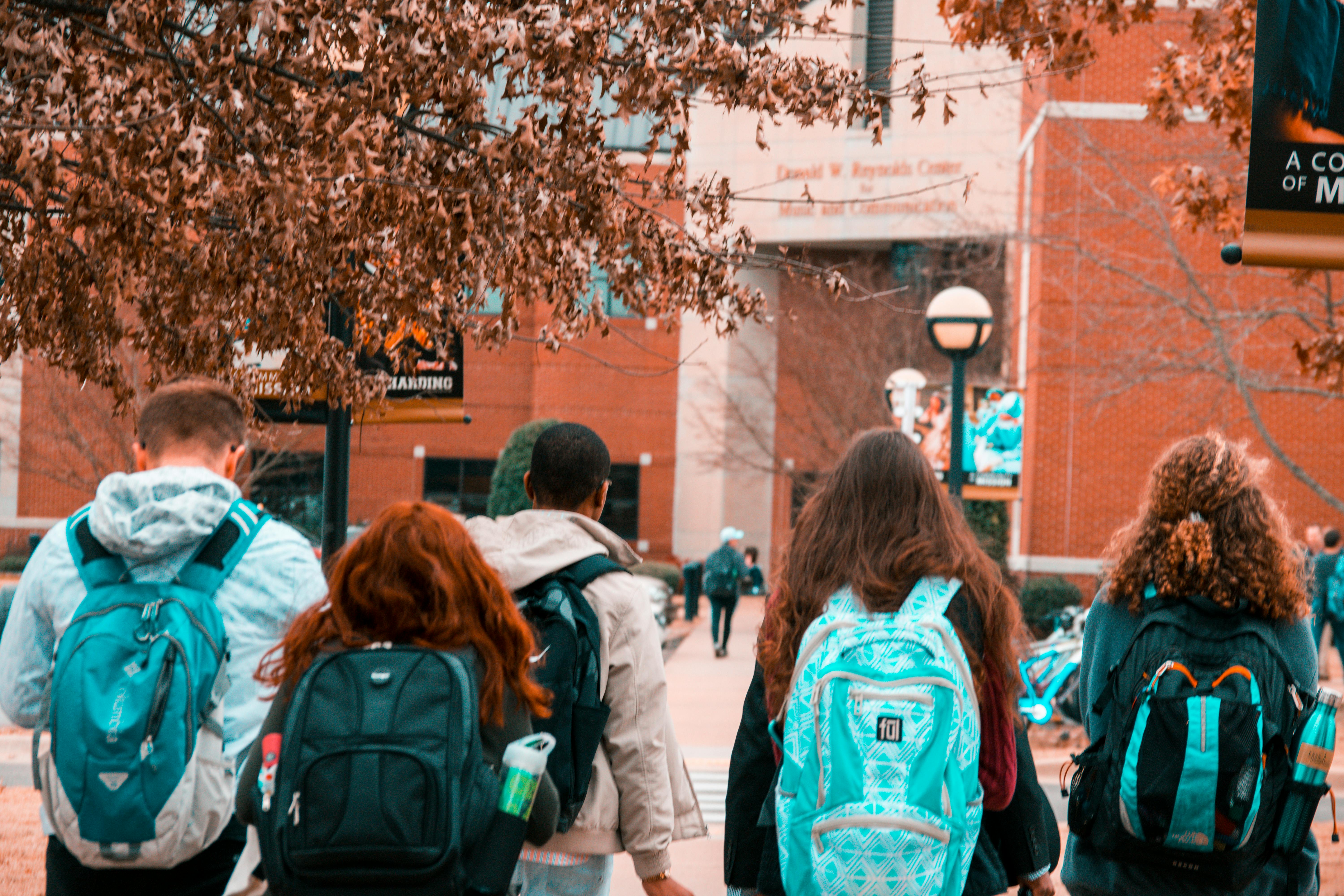 A group of students wearing backpacks walking together on a college campus with fall leaves and brick buildings in the background.