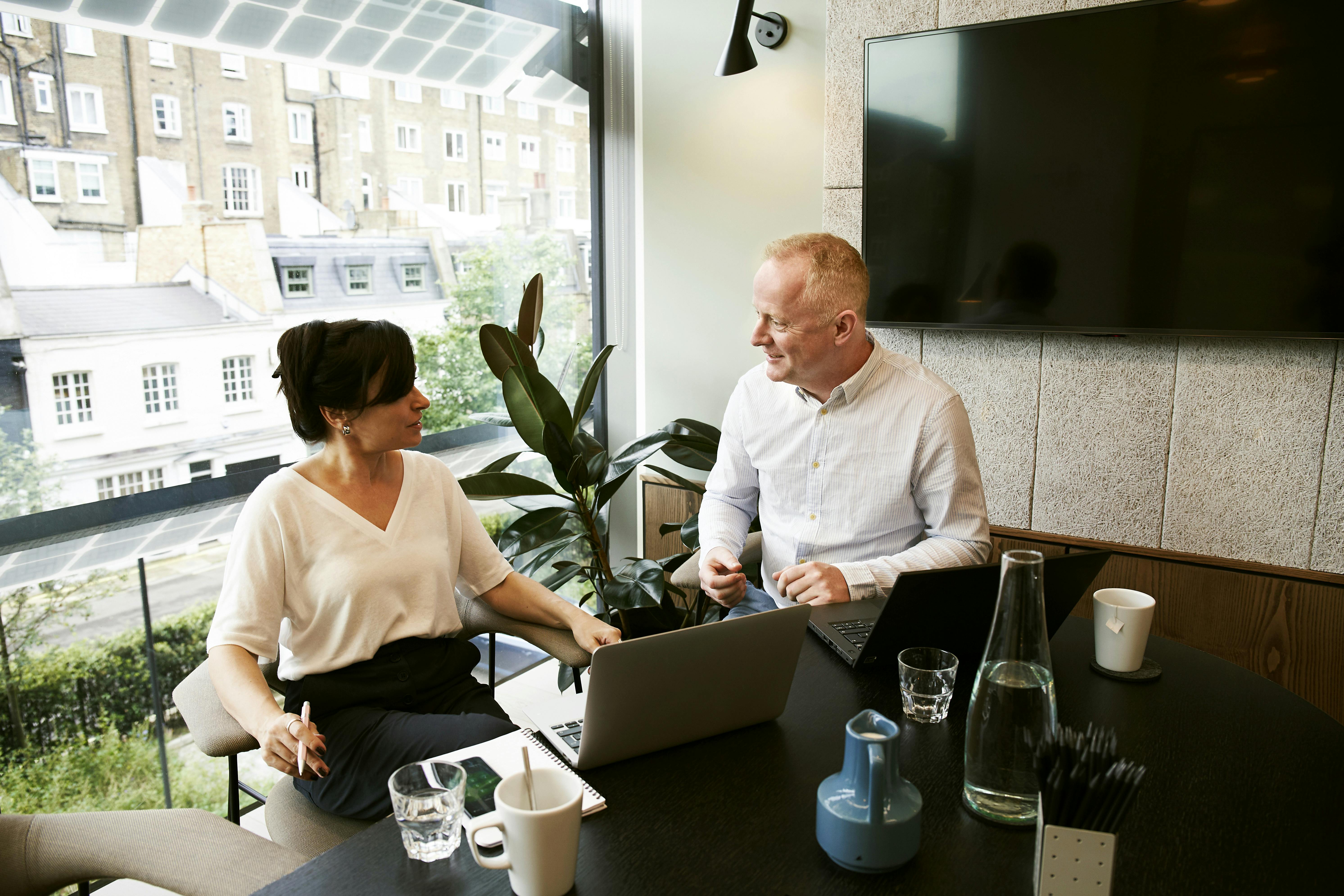 Two business professionals having a discussion at a modern office table with laptops, notebooks, and water glasses near a large window overlooking city buildings.
