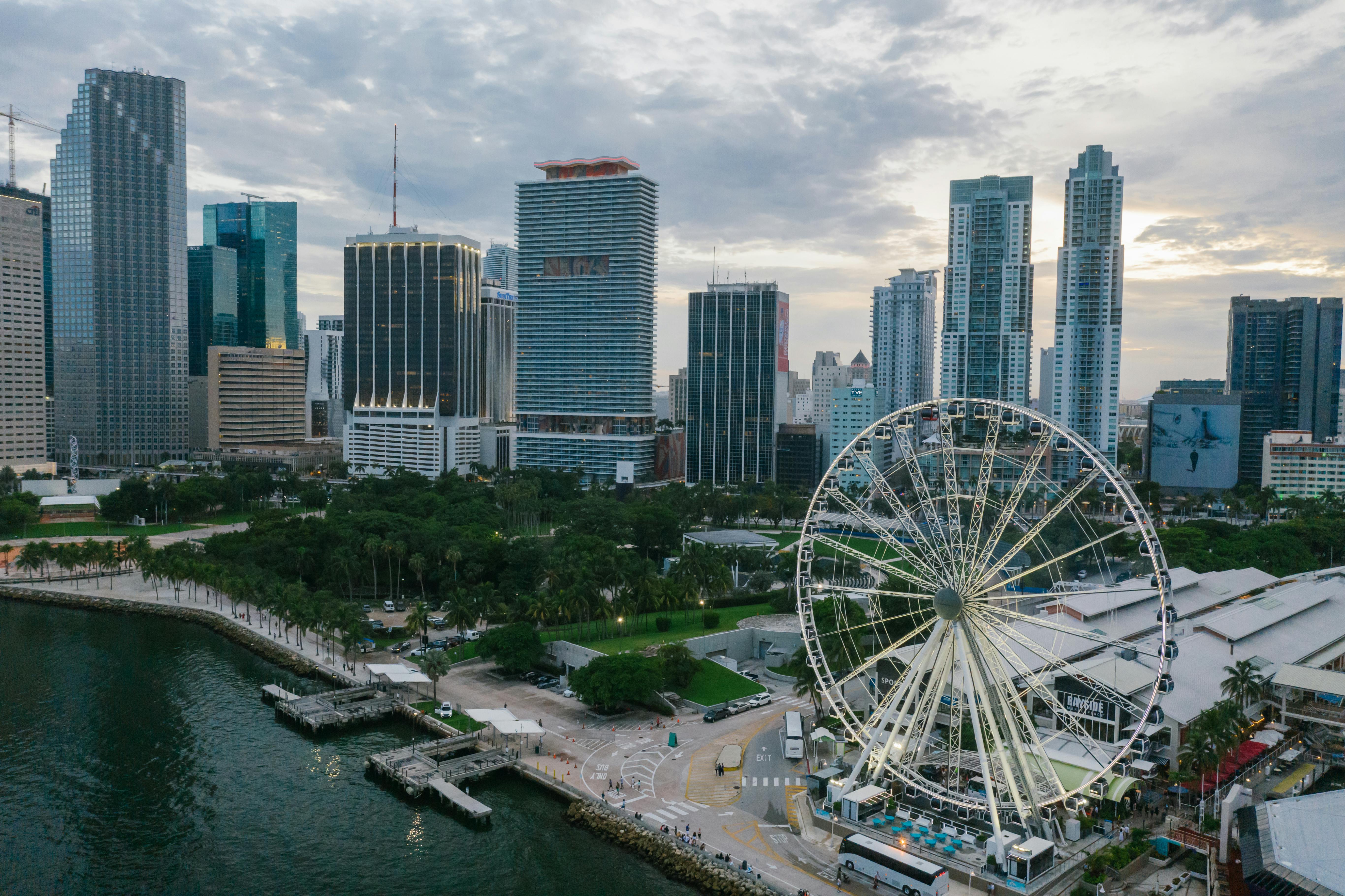 Aerial view of downtown Miami featuring the Skyviews Miami Observation Wheel near Bayside Marketplace, surrounded by modern skyscrapers and Biscayne Bay under a cloudy sky.