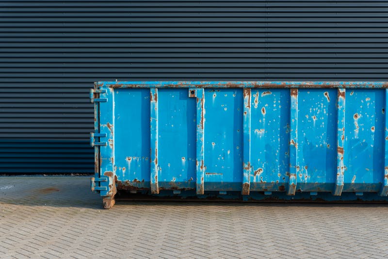 A large, weathered blue dumpster sitting on a paved surface in front of a dark metal wall.
