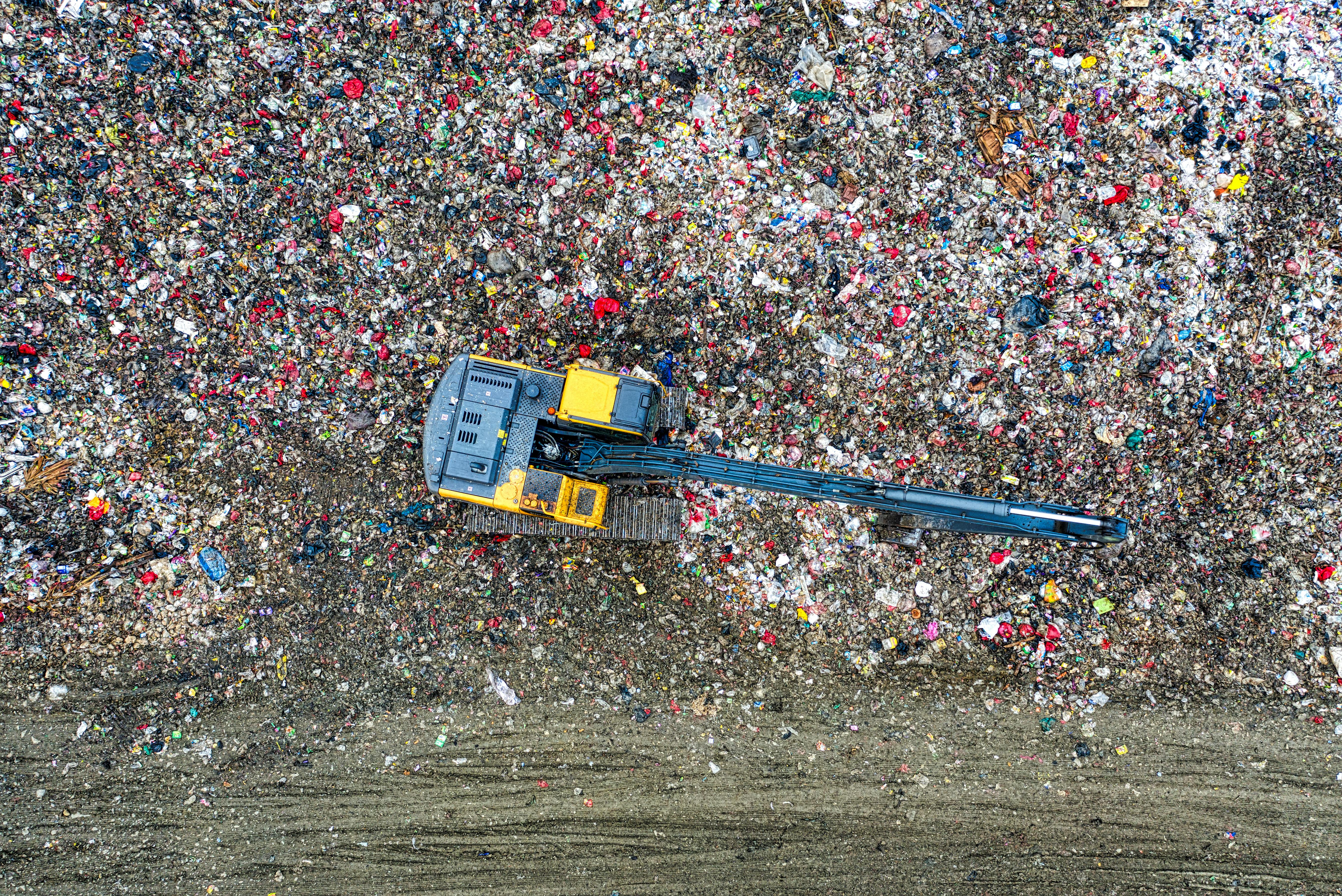 Aerial view of a yellow excavator working on a large landfill covered with mixed trash and debris.