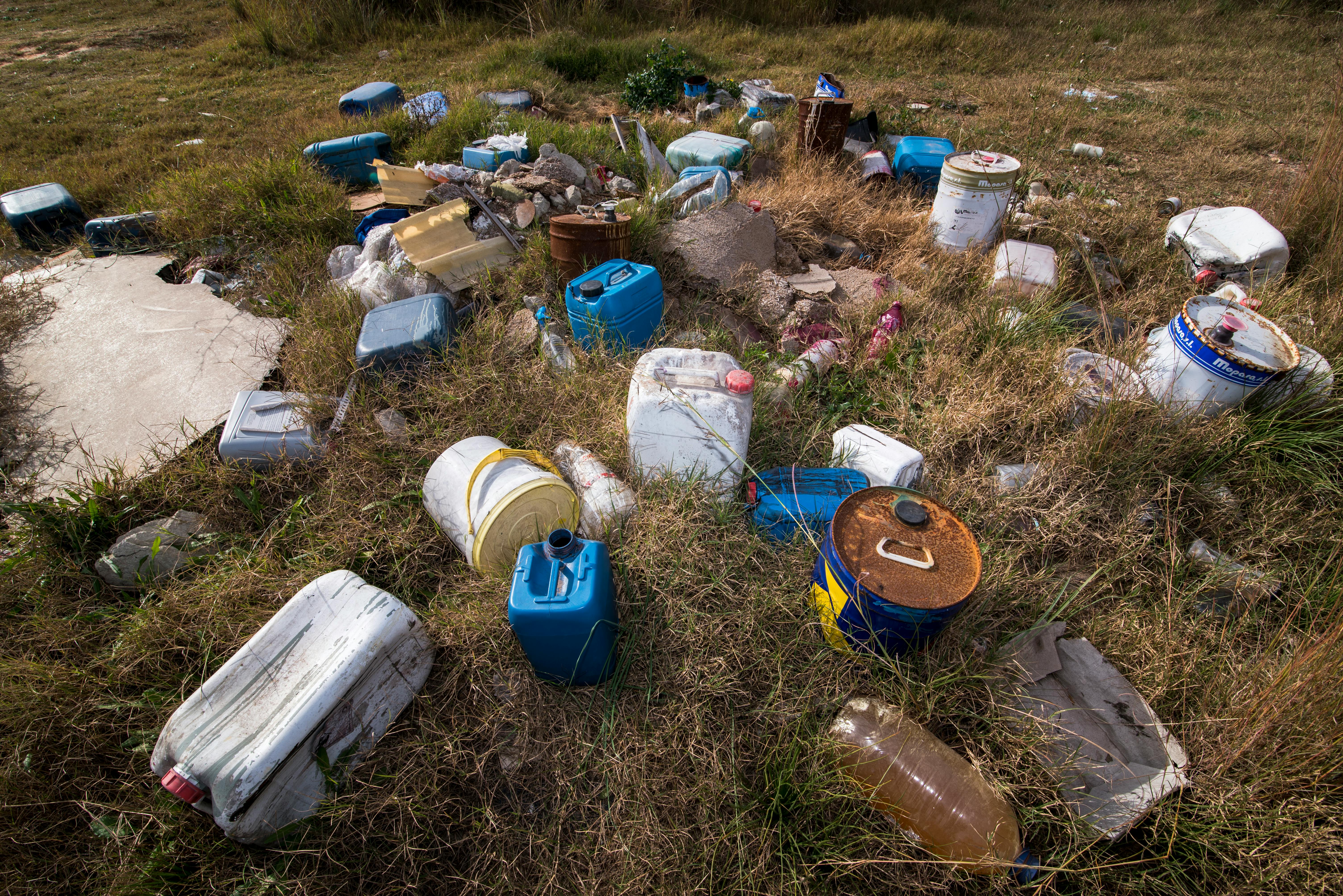 Discarded plastic containers and rusted barrels scattered across a grassy field.