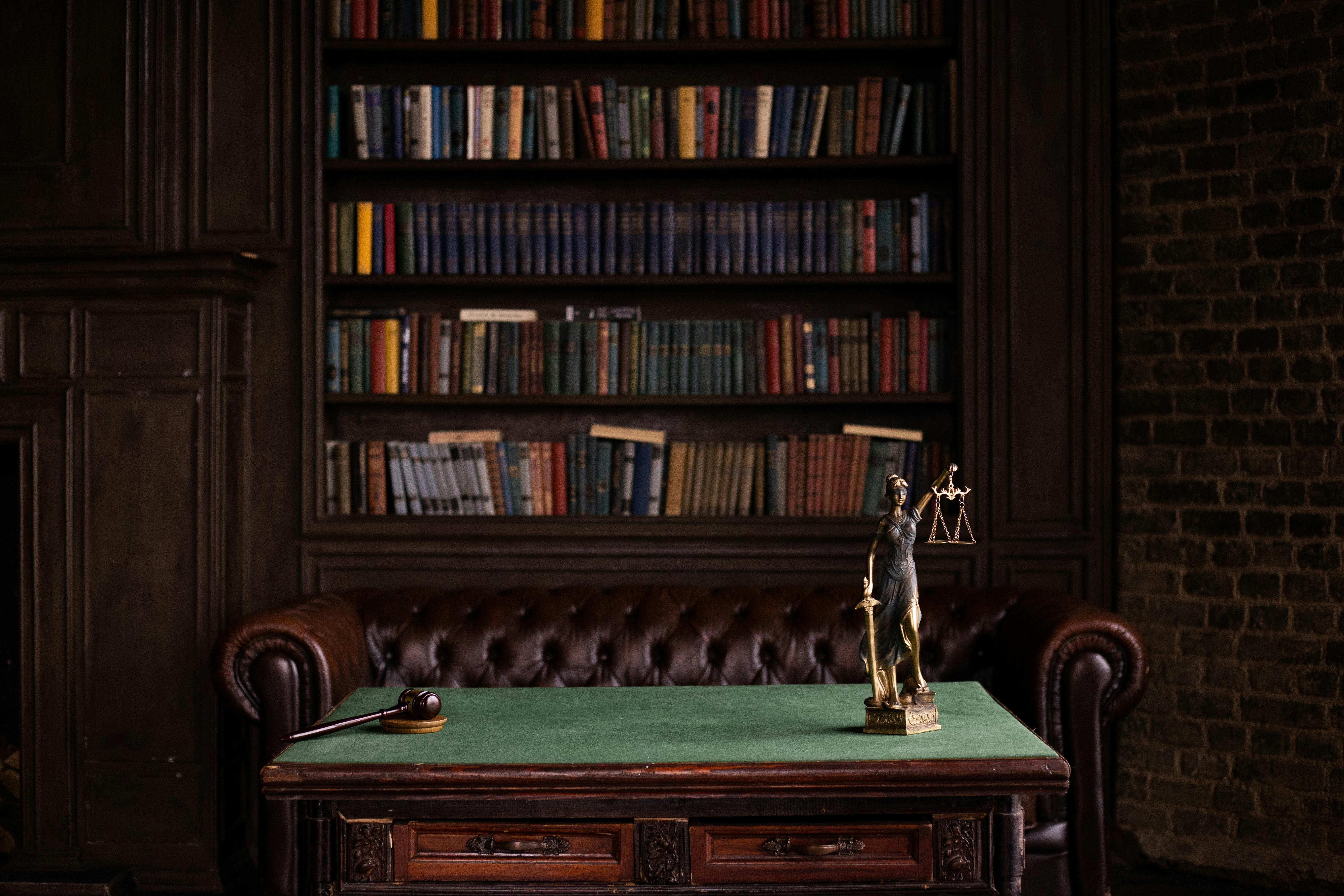 A law office desk with a gavel and a Lady Justice statue in front of a leather sofa and a wall of bookshelves.