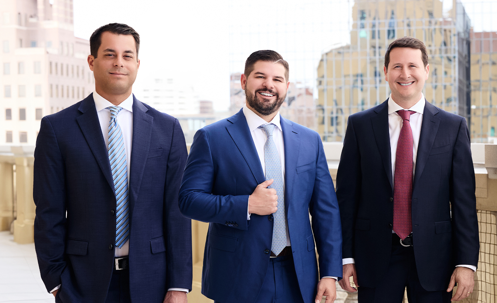 Partners of Vargas Gonzalez Delombard LLP, Rene Delombard (left), Louis Gonzalez (middle), and Andrew Vargas (right) - standing outdoors with a cityscape in the background, dressed in business suits and smiling