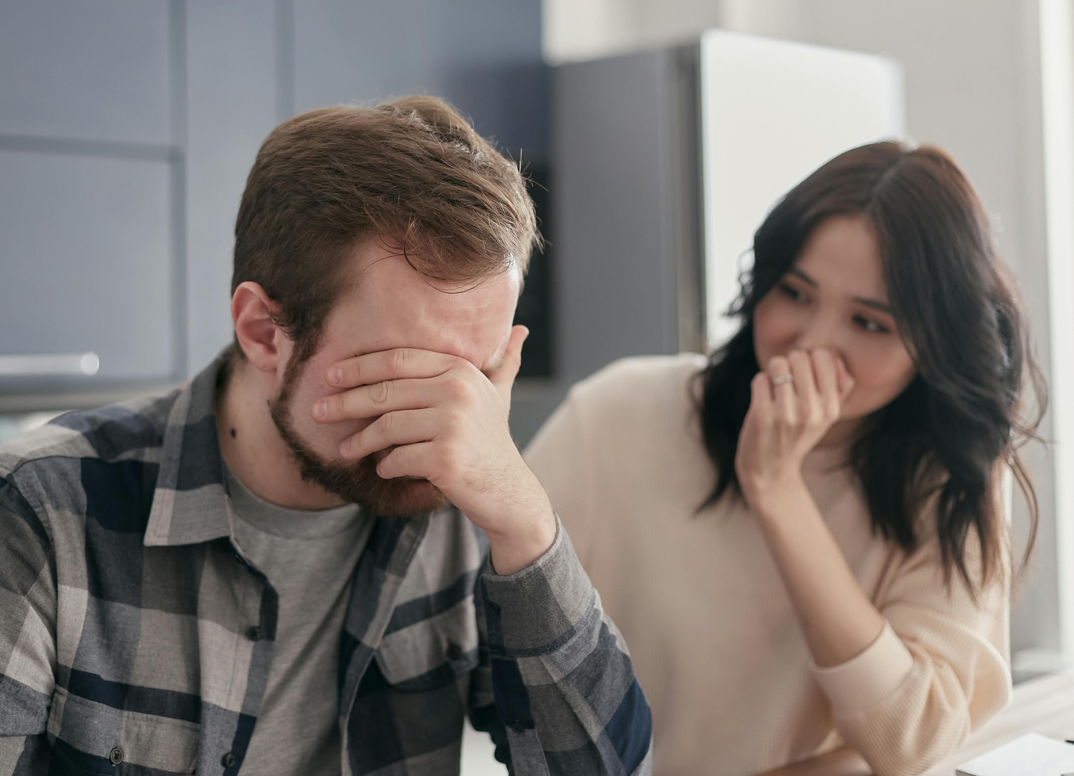 A man sits with his face in his hands, looking stressed or overwhelmed, while a woman beside him gently covers her mouth with her hand, appearing concerned. 