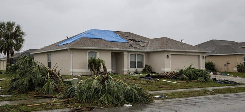 Home with severe roof damage and a blue tarp covering part of the roof after a hurricane, with fallen palm trees and debris scattered across the yard