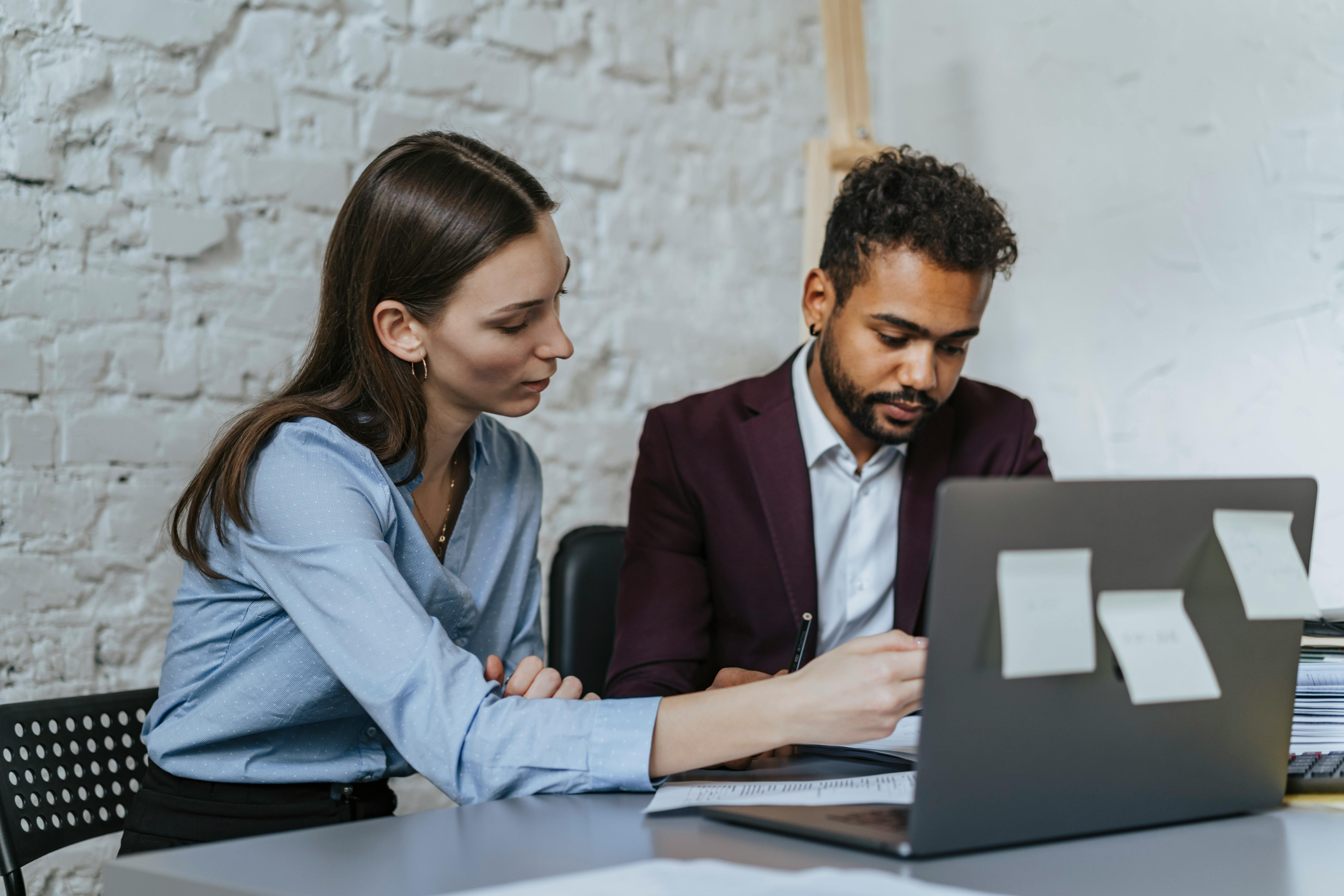 Two colleagues reviewing documents together at a desk, looking at a laptop with sticky notes on it