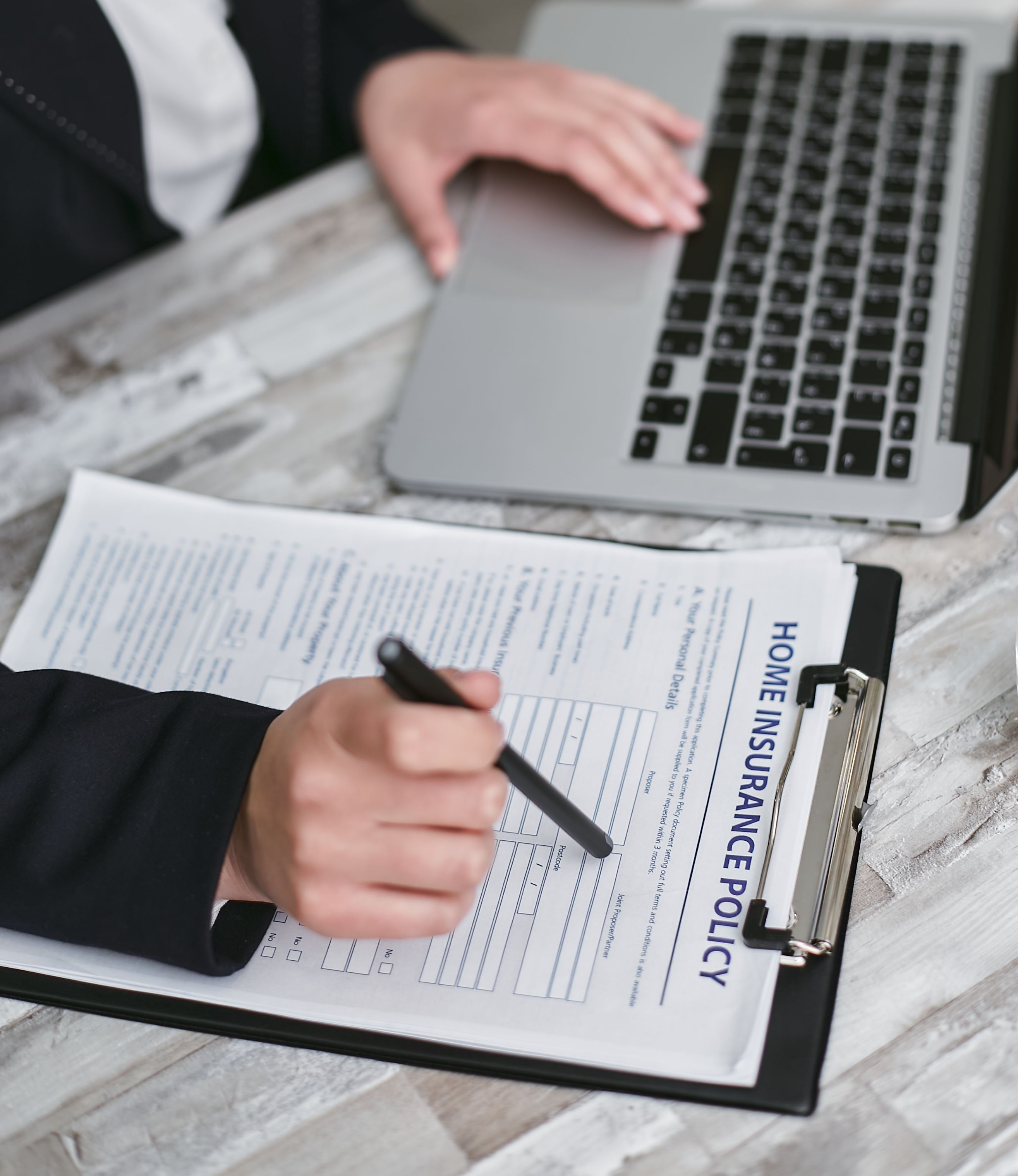 Person reviewing a home insurance policy on a clipboard while using a laptop to prepare for a property damage insurance claim.