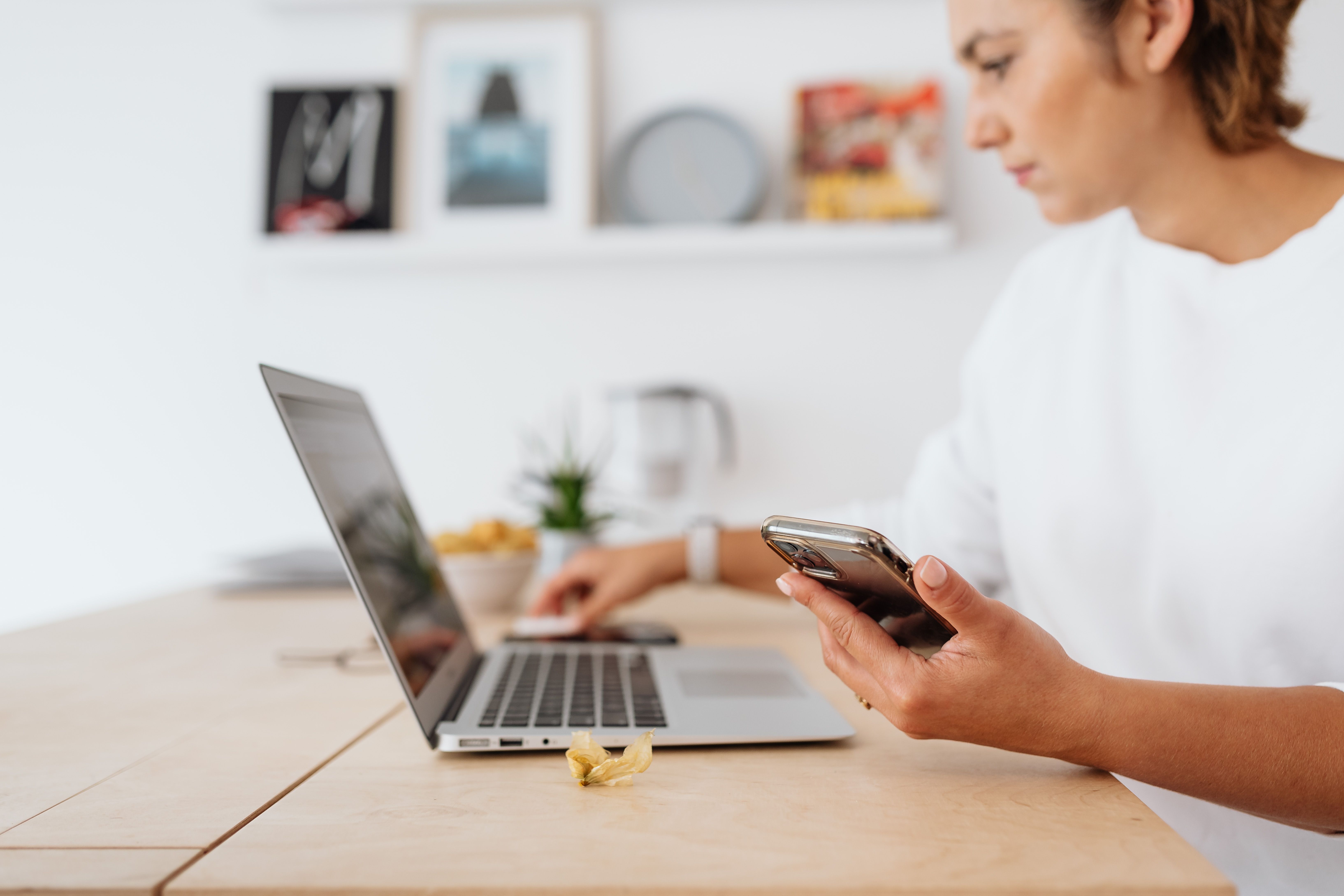 Woman using a laptop and holding a phone while sitting at a table.