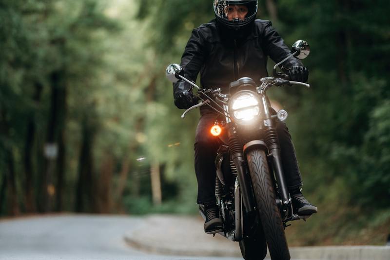 Motorcyclist wearing black protective gear riding a motorcycle with its headlight on along a tree-lined road.