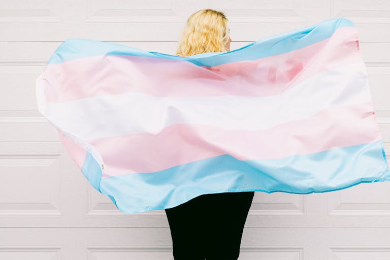 Transgender person from behind, wearing pink and white striped sweatshirt, holds transgender flag