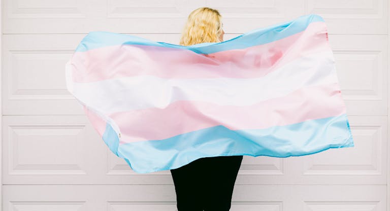 Transgender person from behind, wearing pink and white striped sweatshirt, holds transgender flag