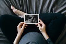 High angle shot of Asian pregnant woman holding an ultrasound scan photo in front of her baby bump, sitting on bed at home. Mother-to-be. Precious moment in life. Preparation for a new family member. Expecting a new life. Baby and new life concept