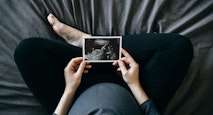 High angle shot of Asian pregnant woman holding an ultrasound scan photo in front of her baby bump, sitting on bed at home. Mother-to-be. Precious moment in life. Preparation for a new family member. Expecting a new life. Baby and new life concept