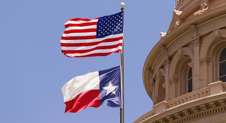 The Texas flag and US flag fly in front of Texas capitol building.