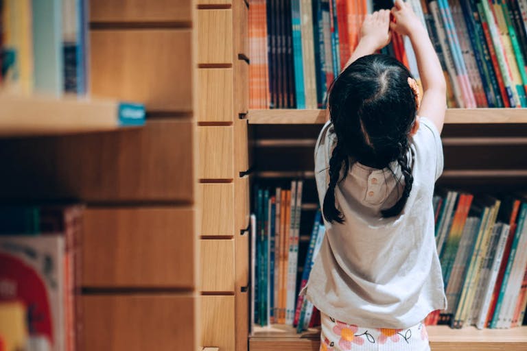 Little Asian girl choosing books from the bookshelf in library How a Planned Parenthood coalition fights to keep sexually graphic books in schools featured image