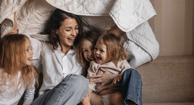 Happy mother with three girls holding blanket on sofa at home
