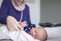 Midwife examining newborn sleeping baby girl with stethoscope on bed at home