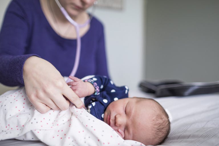 Midwife examining newborn sleeping baby girl with stethoscope on bed at home