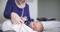 Midwife examining newborn sleeping baby girl with stethoscope on bed at home